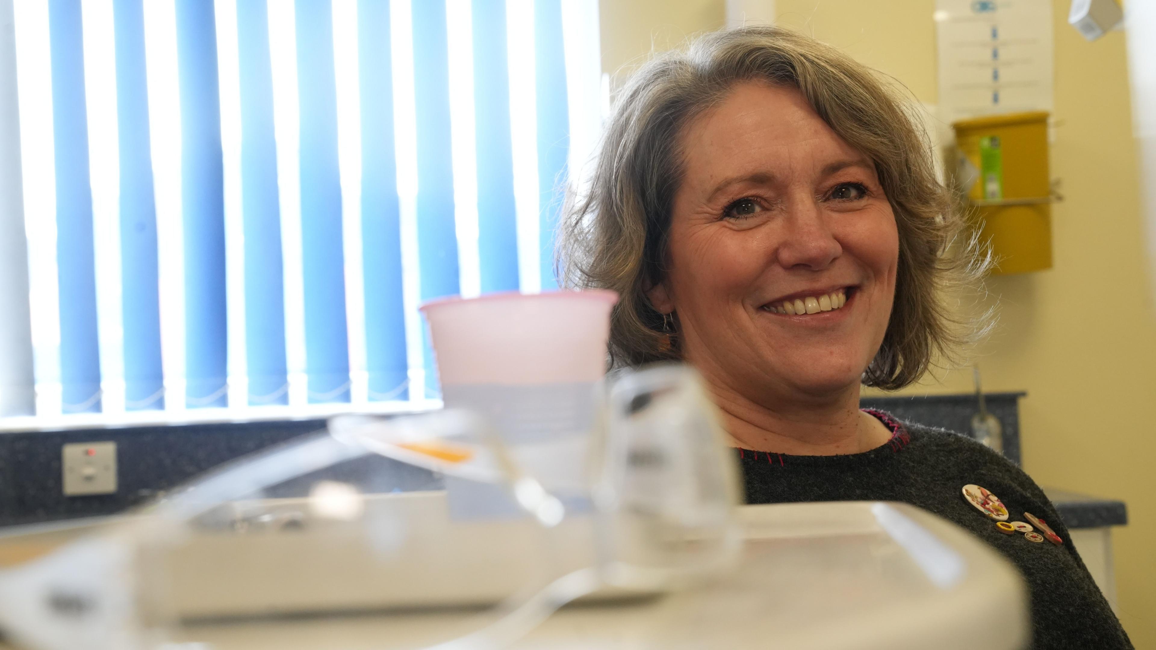 Patient Caroline Poole sits in the dentist's chair with her head and shoulders visible. She is wearing a grey jumper with an assortment of buttons on the left hand side. Caroline is smiling and in the foreground of the image is a pair of dental glasses and a cup of mouthwash, which is slightly out of focus.