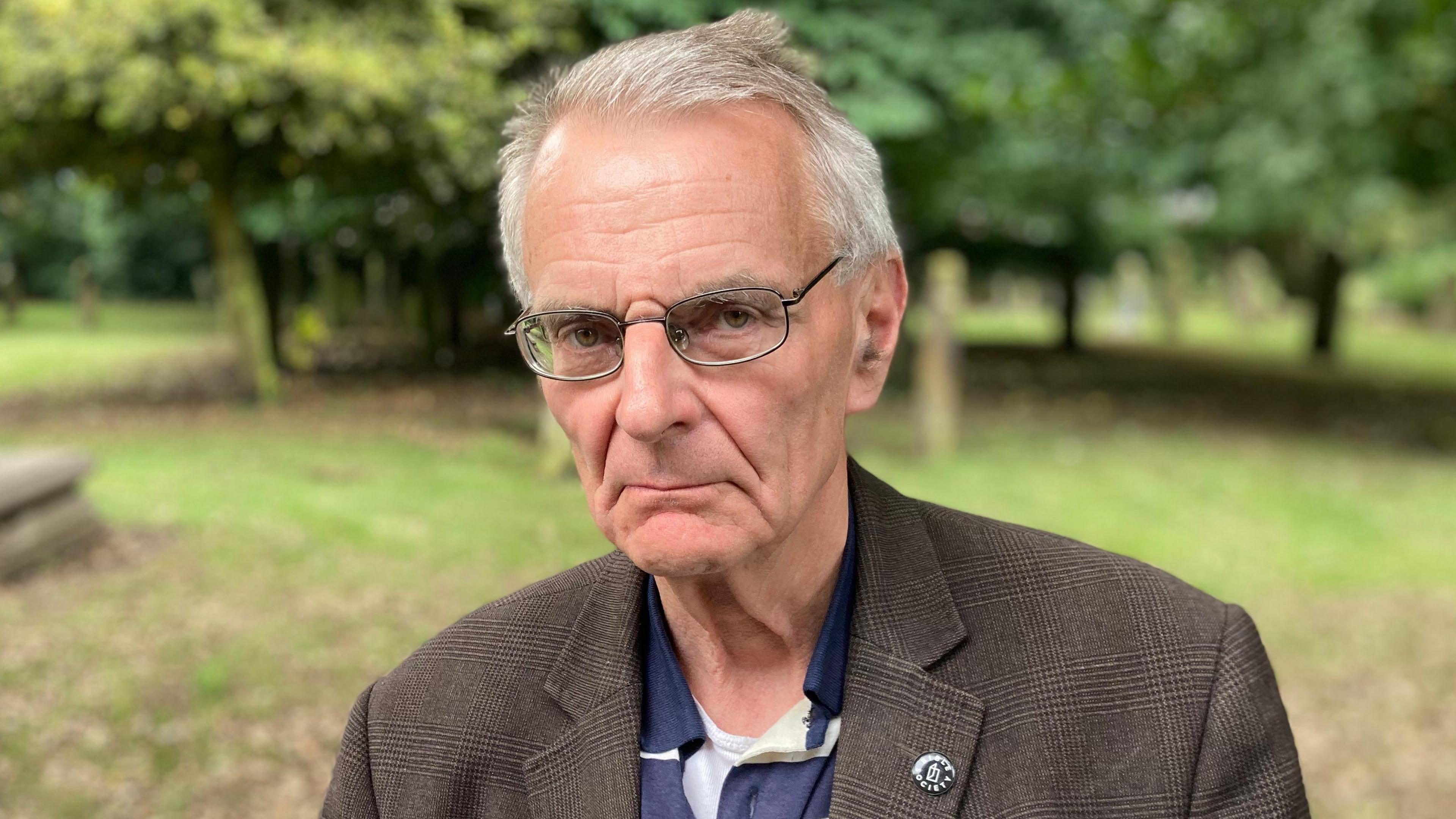Charles Wellington is wearing glasses and a tweed jacket. He has a sombre expression on his face.