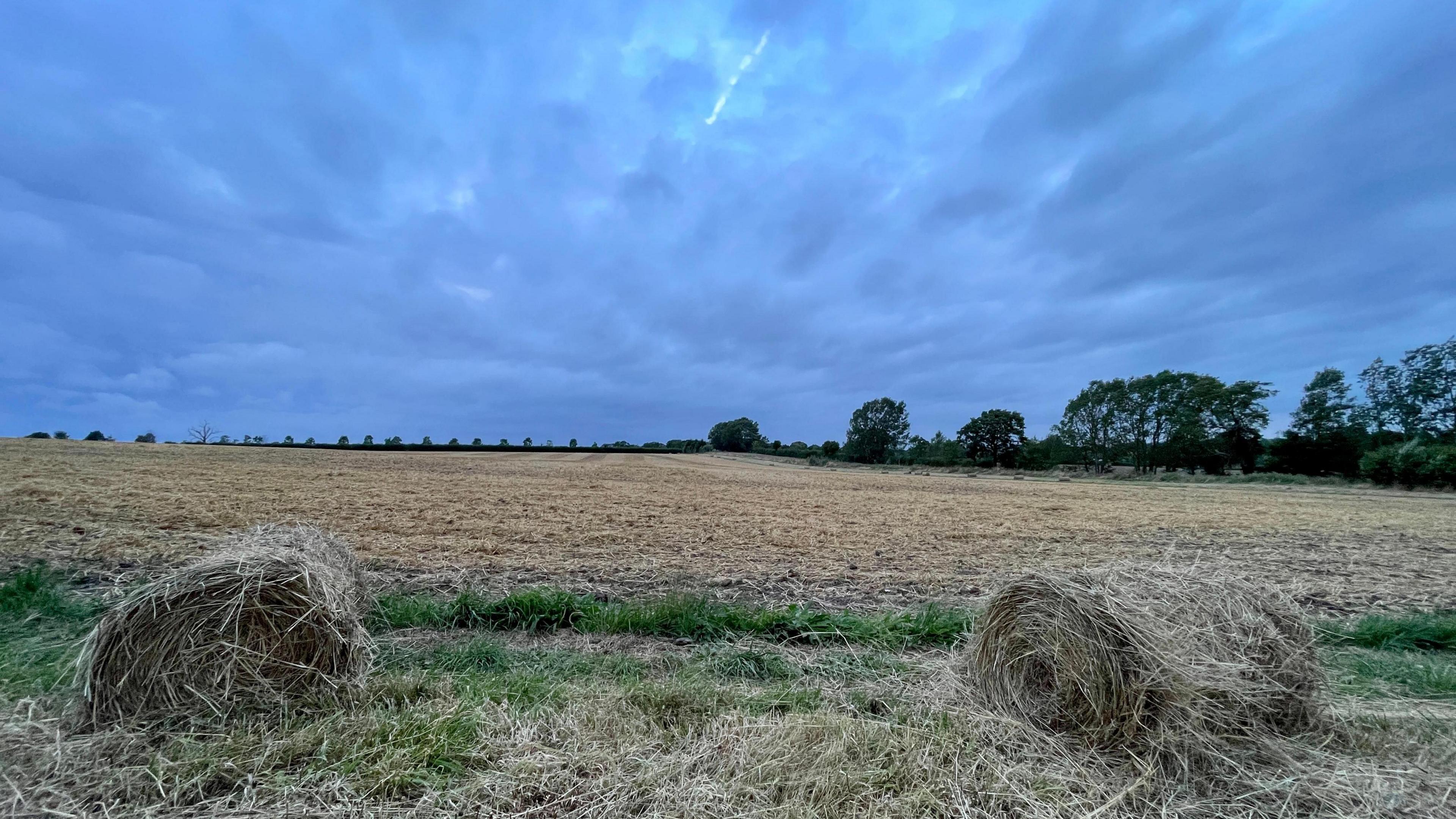 A stormy dark grey sky full of clouds over a field of cut crops near Binfield. There are two round bales that look depressing and are falling apart on a patch of grass. On the horizon is a row of trees. 