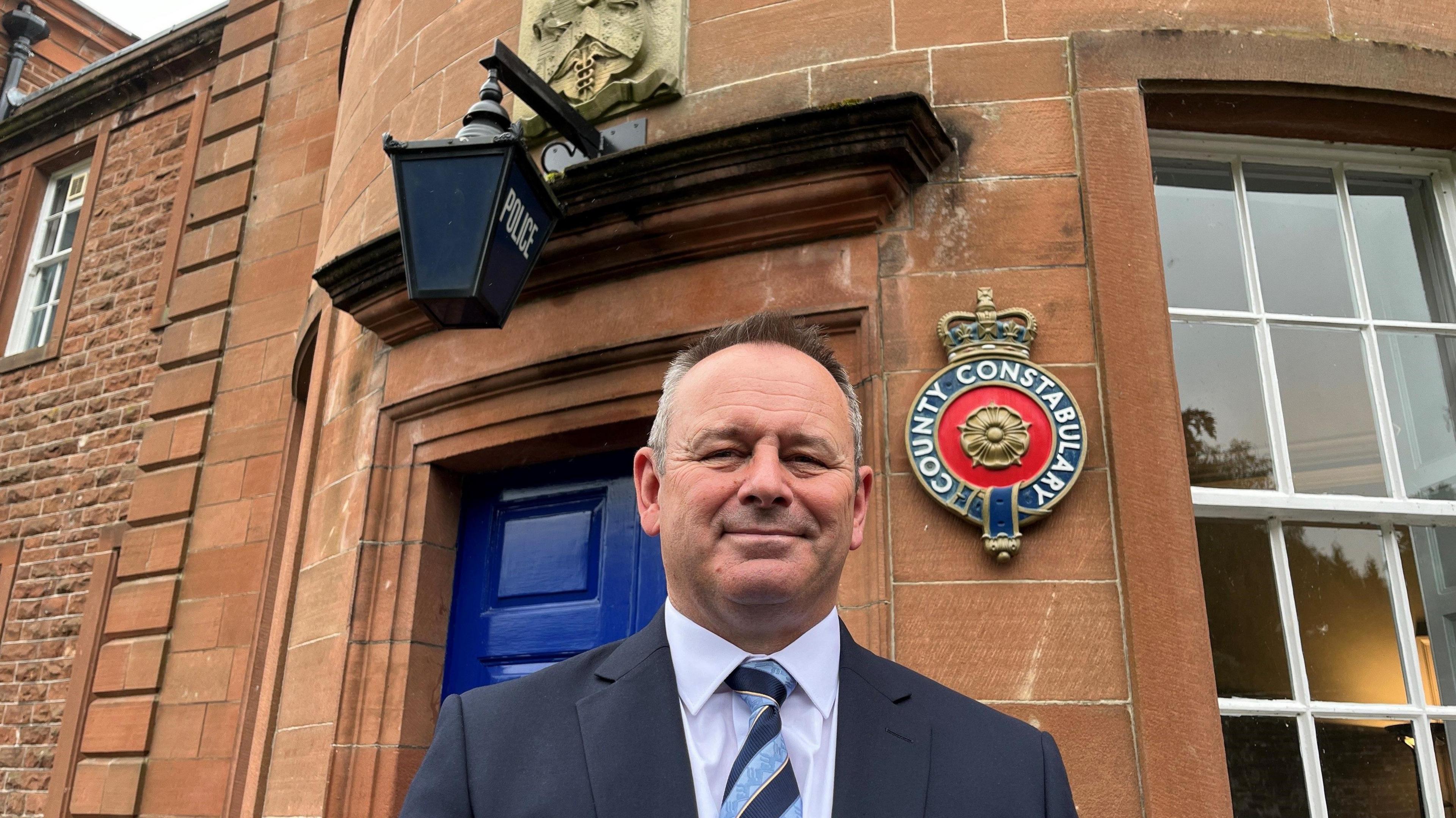 Head and shoulder view of David Allen, the new Cumbria Police Fire and Crime Commissioner, wearing a suit and tie and standing in front of a doorway with a blue police lantern above it