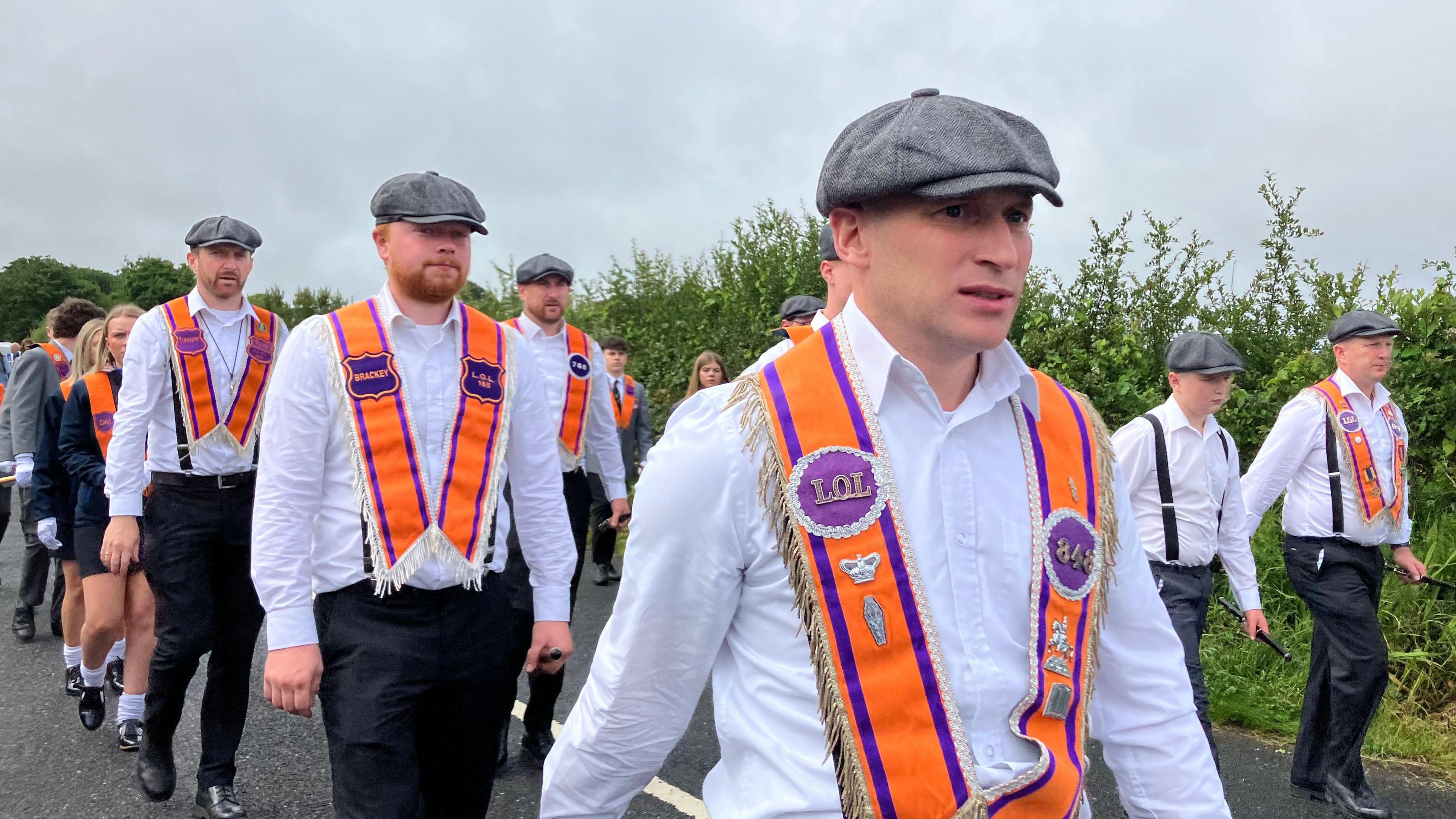 Men marching in white shirts and with orange sashes around their shoulders and grey flat caps
