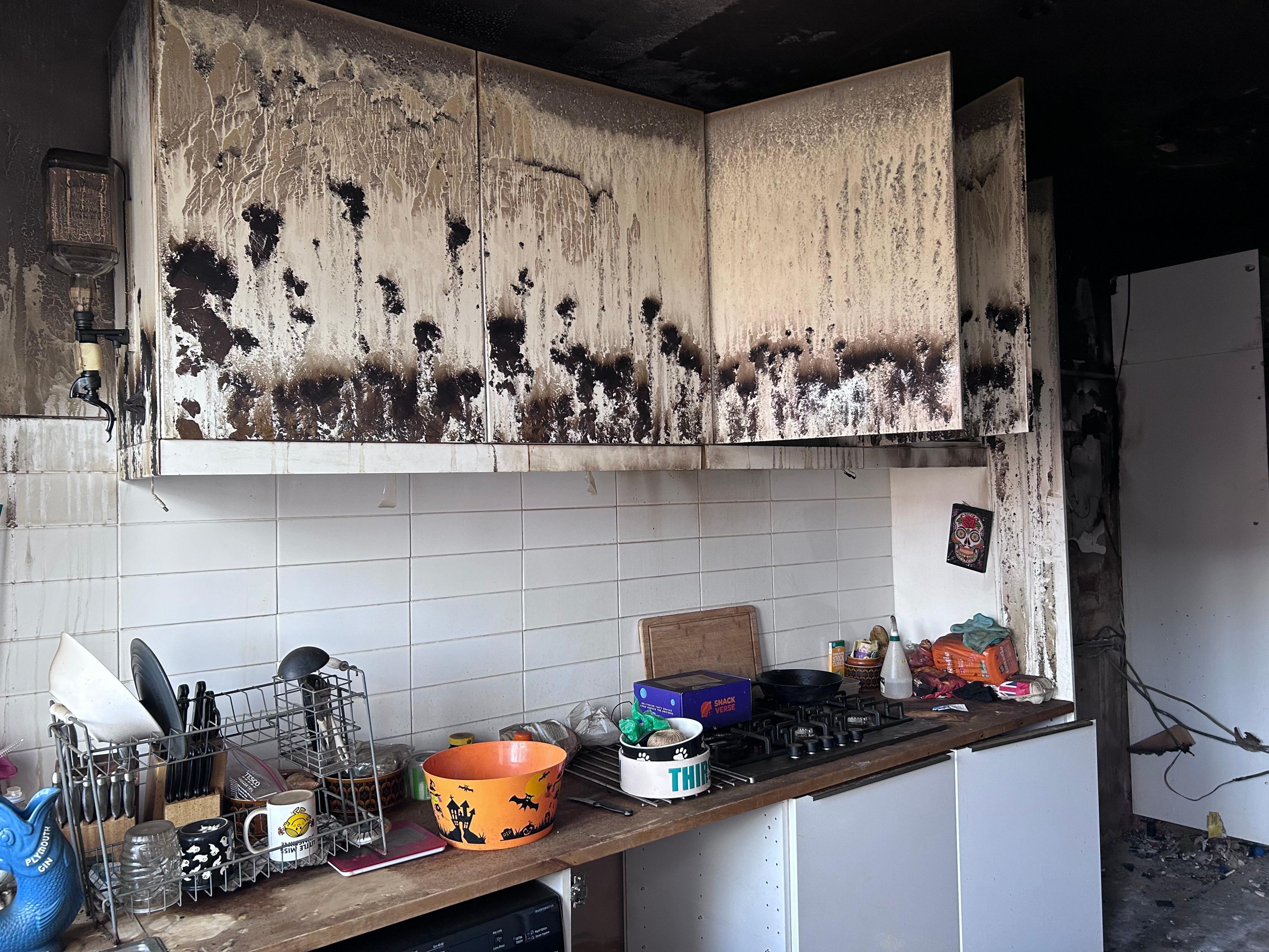 A burnt out kitchen where the units have warped and blackened due to heat and smoke damage. There are items on the kitchen bench and on the drainer.