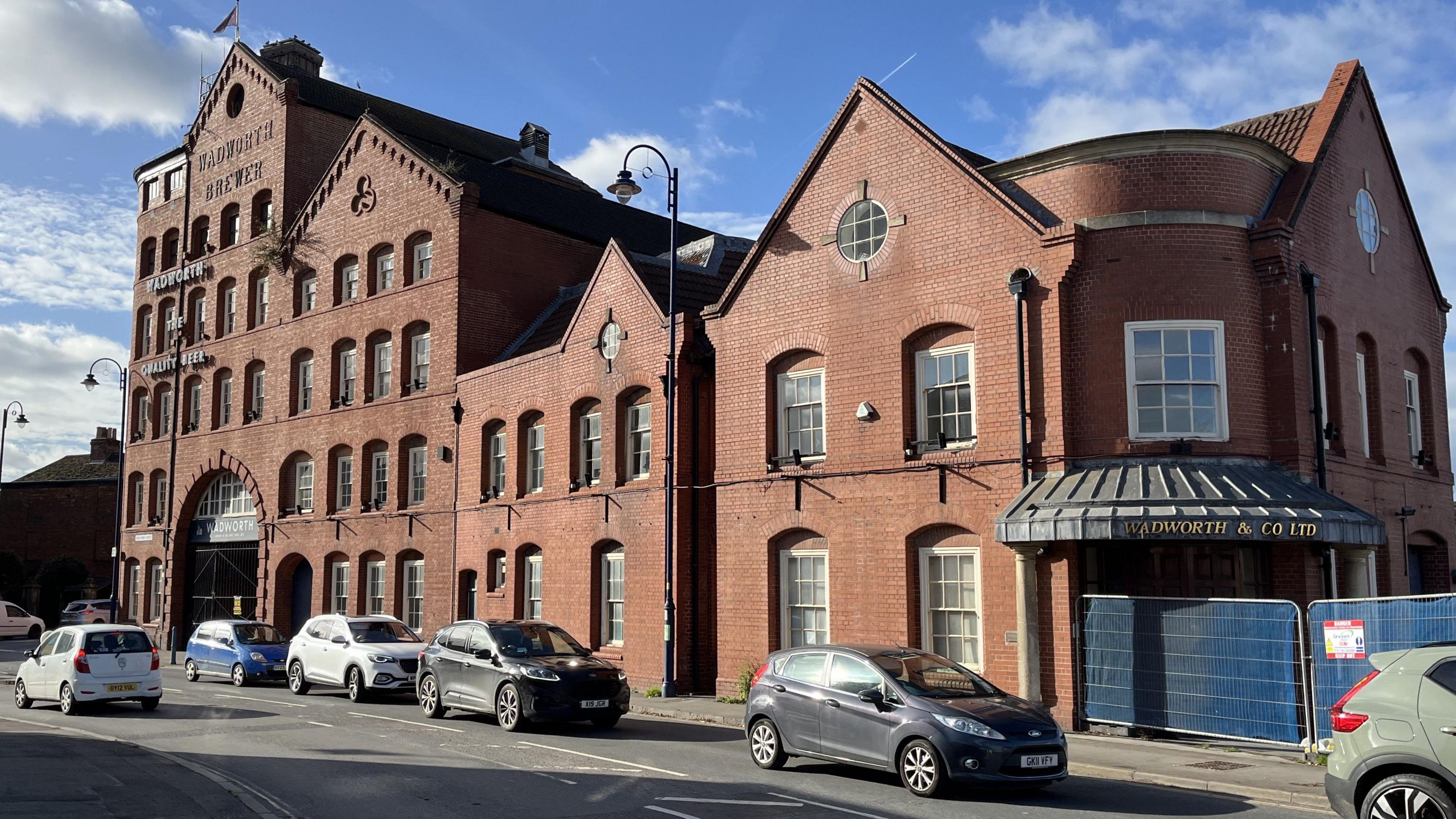 Cars pass the former brewery buildings of Wadworth & Co in Devizes. The main building of the red brick Victorian warehouse-style brewery is seven storey's high with a selection of similar red brick buildings standing next to it. 
Signs saying Wadworth can still be seen. 
