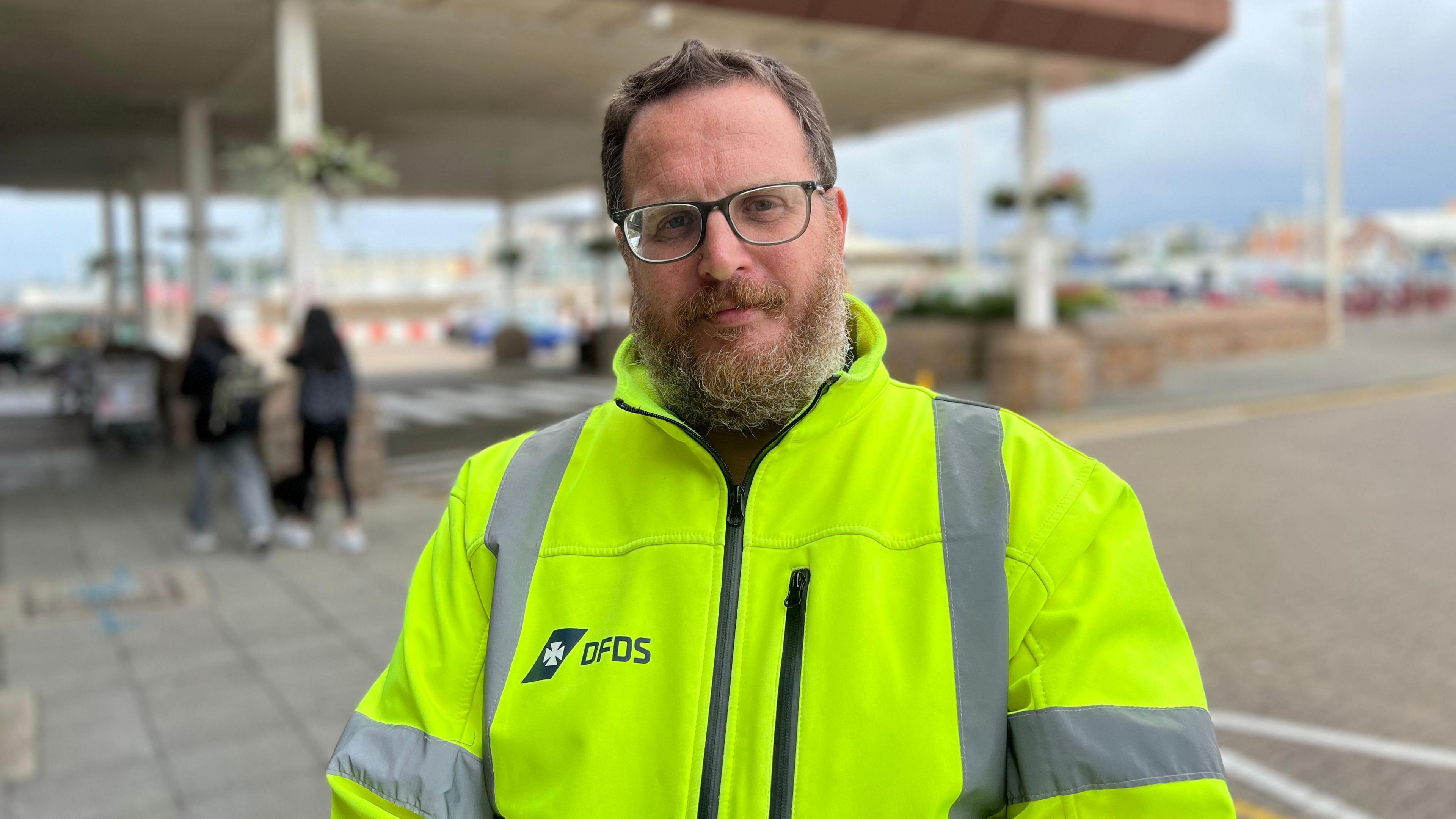 A man wearing a high vis jacket with DFDS logo on the right chest area, background of the harbour is blurred.