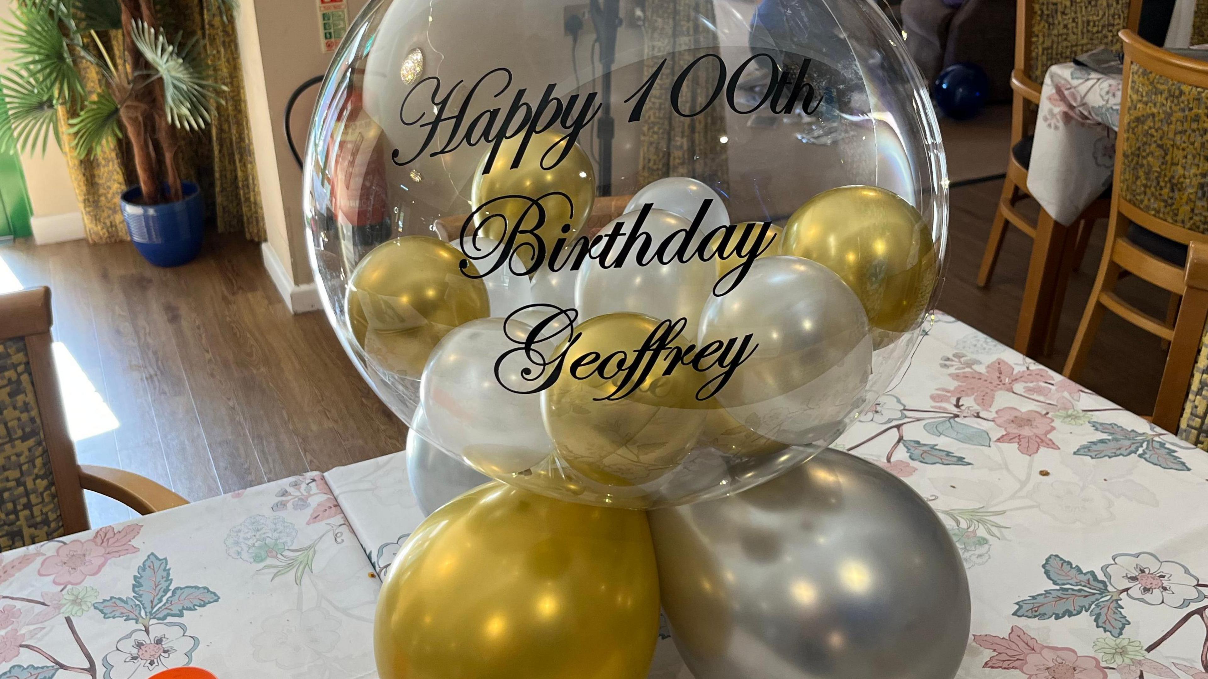 A large clear balloon on a table with smaller gold and white balloons inside and at its base. In black writing on the front it reads 'Happy 100th birthday Geoffrey'