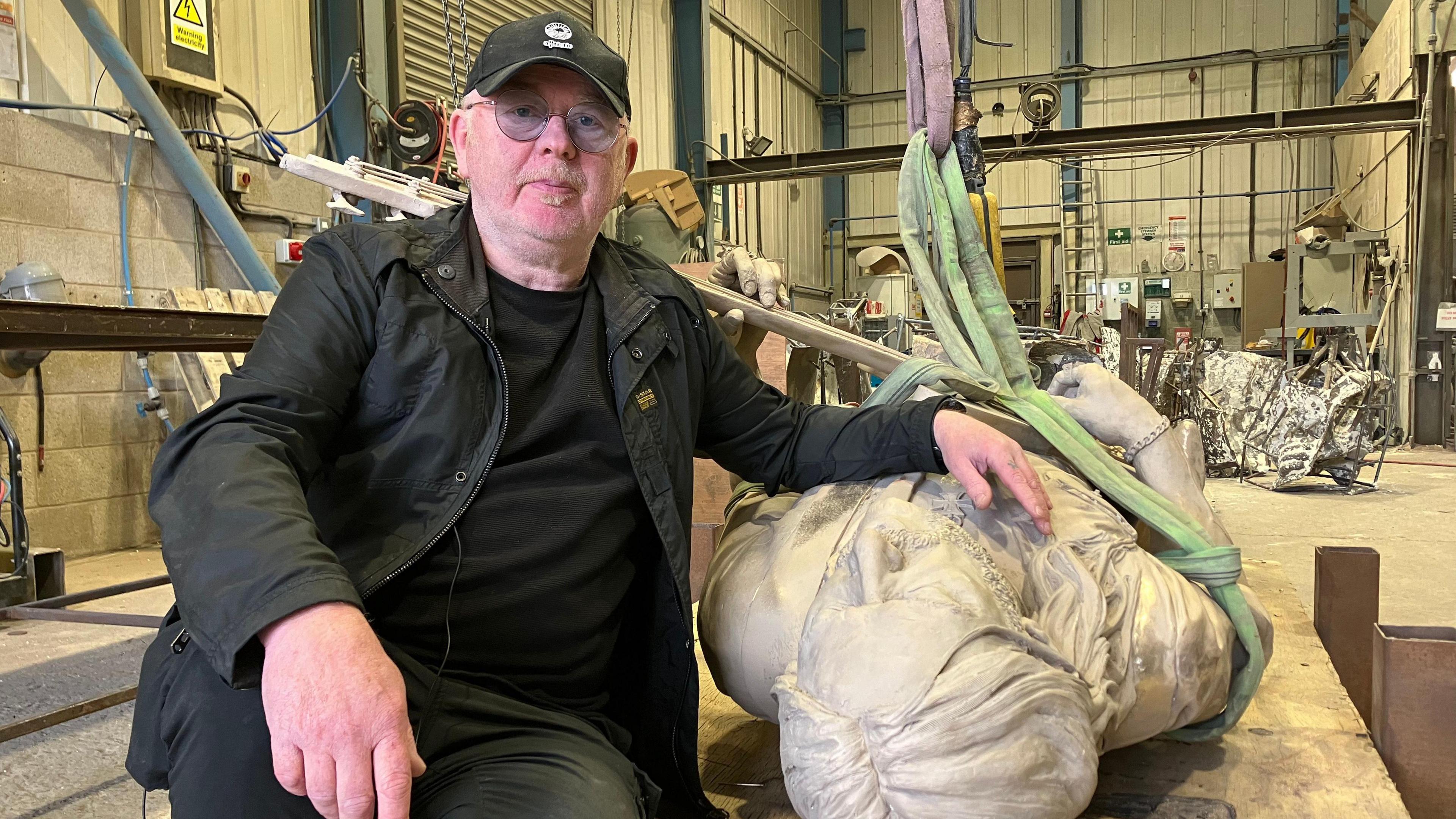 A man with a blonde soul patch and glasses, wearing a black baseball cap, a black t-shirt under a black jacket and black jeans, sits with his hand resting on a statue which is lying on its back.