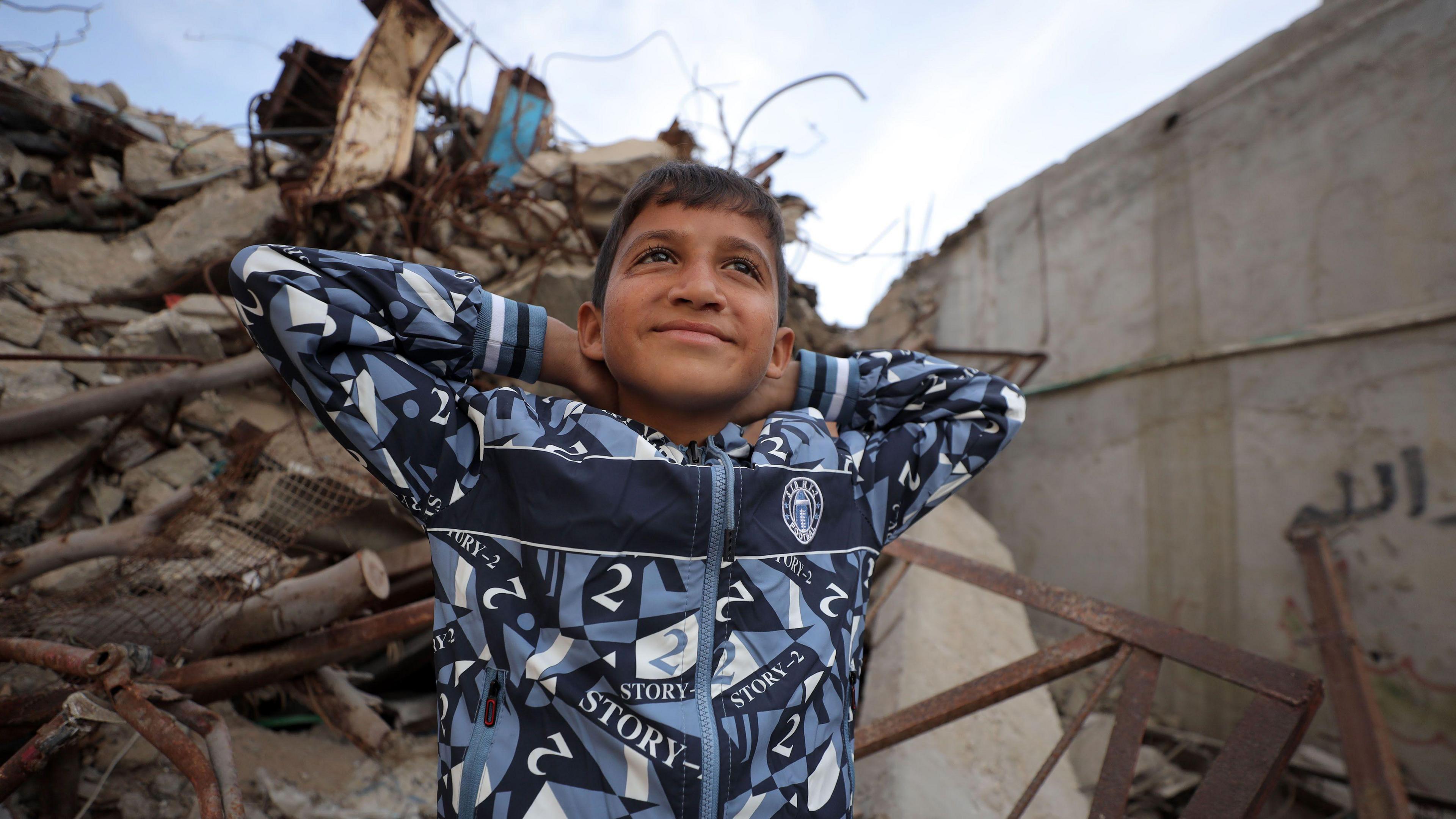 Zakaria wears a blue and white hoodie as he sits on some rubble and puts his hands behind his head while smiling.