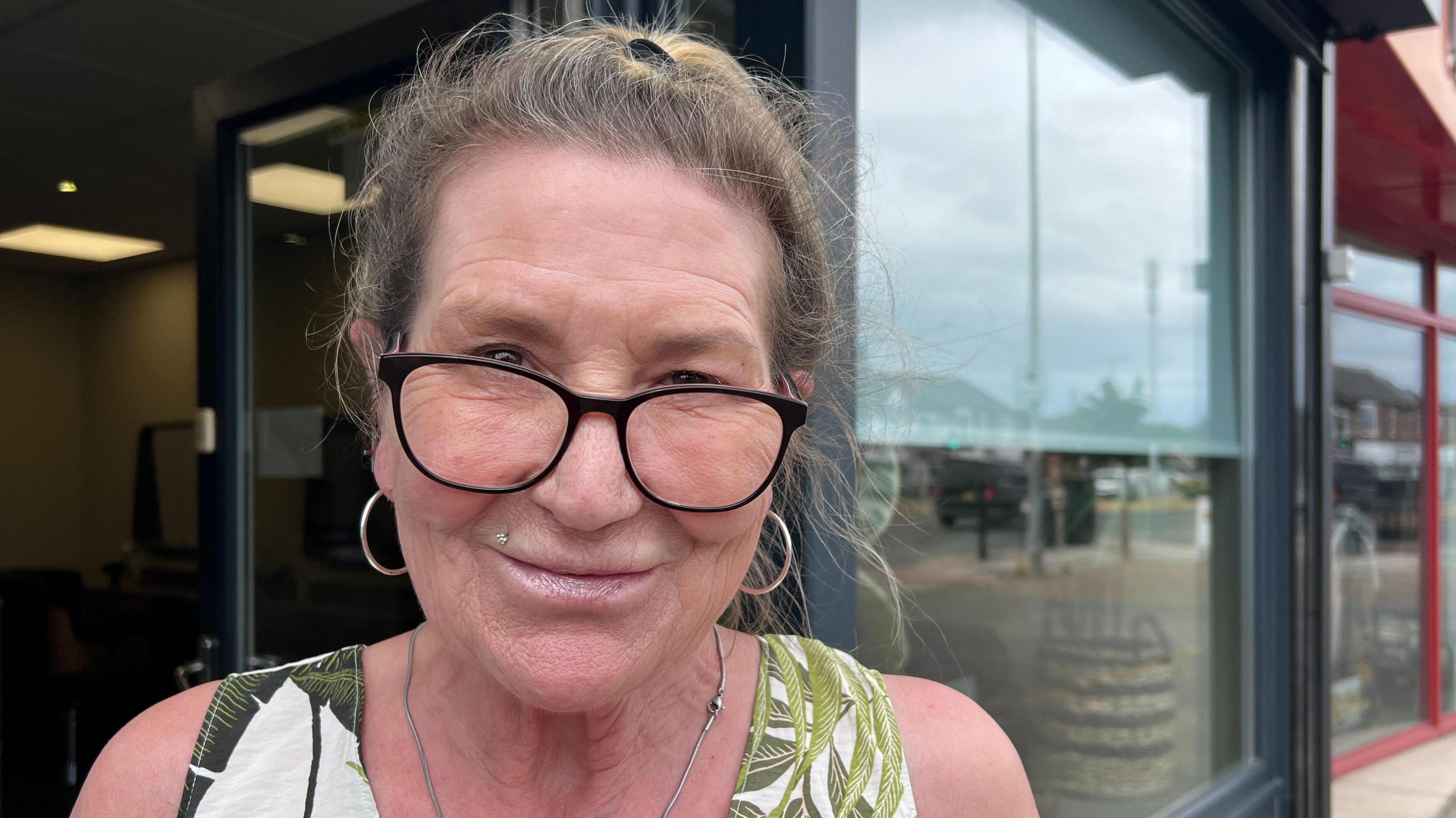 Christine Colville has her hair tied back and is wearing black glasses, gold hooped earrings and a vest top printed with a green botanical design. She is standing in front of a retail outlet with large glass windows.
