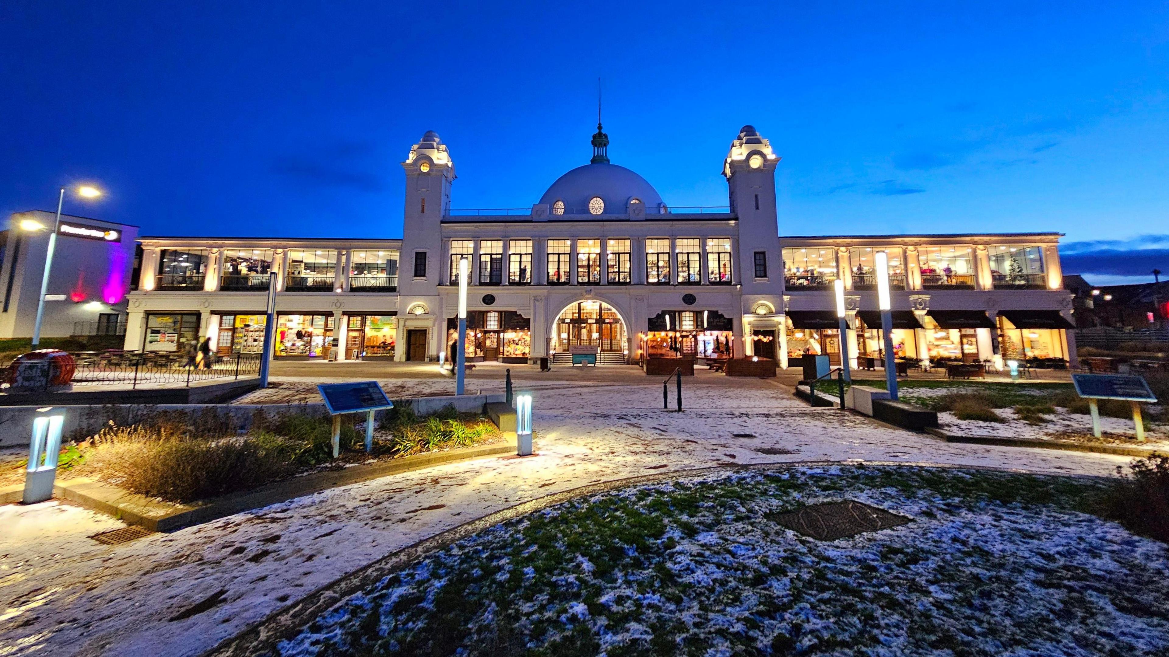 Spanish City is a grand two-storey building made of white concrete. There is a dome on top. All the windows are lit, which contrasts with the dark blue sky at dusk. There is a dusting of snow on the plaza out front.