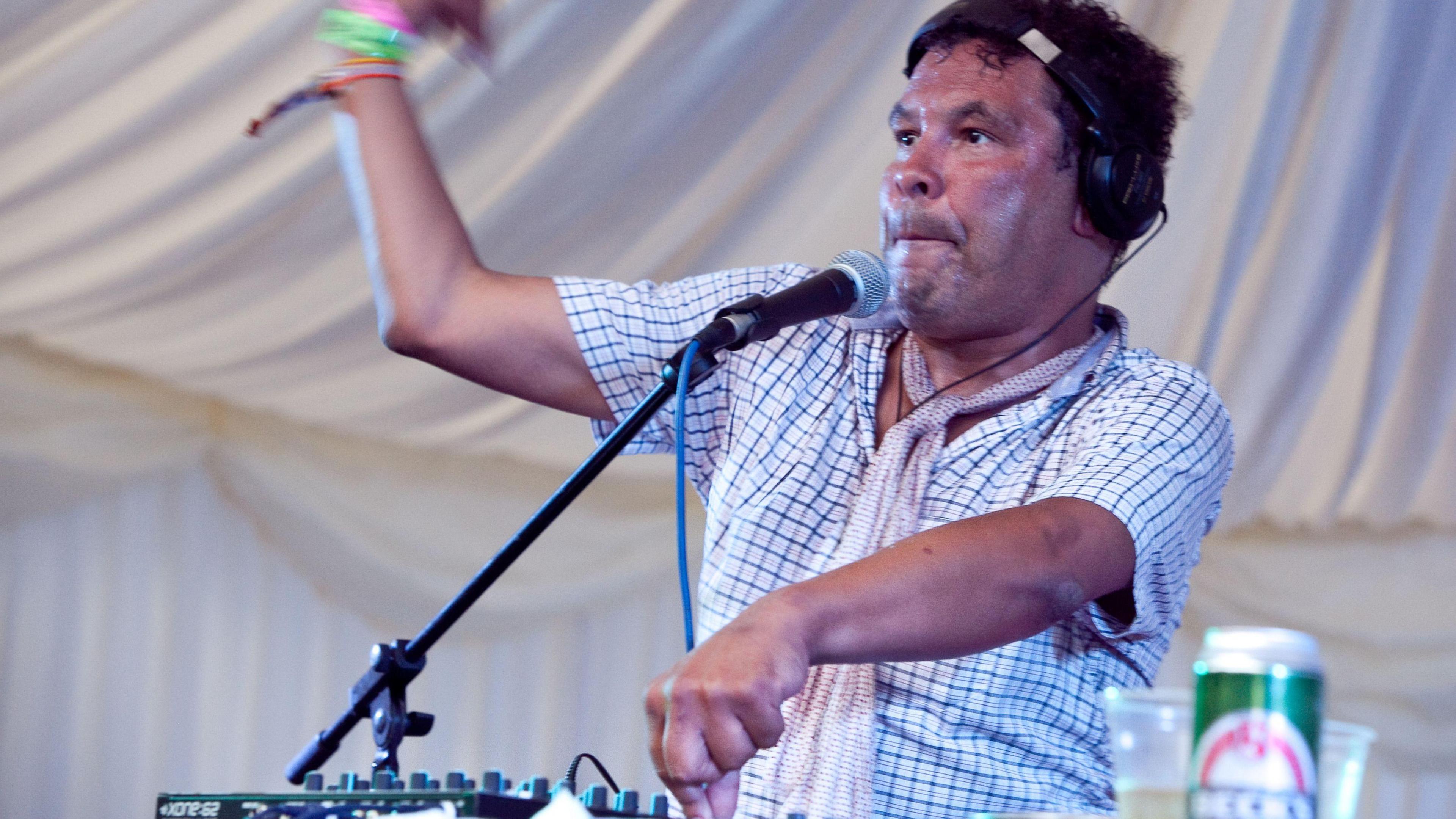 DJ Craig Charles is wearing a check shirt with a thin red scarf around his neck. He is wearing headphones and thumping his hand in the air as he plays a music set.