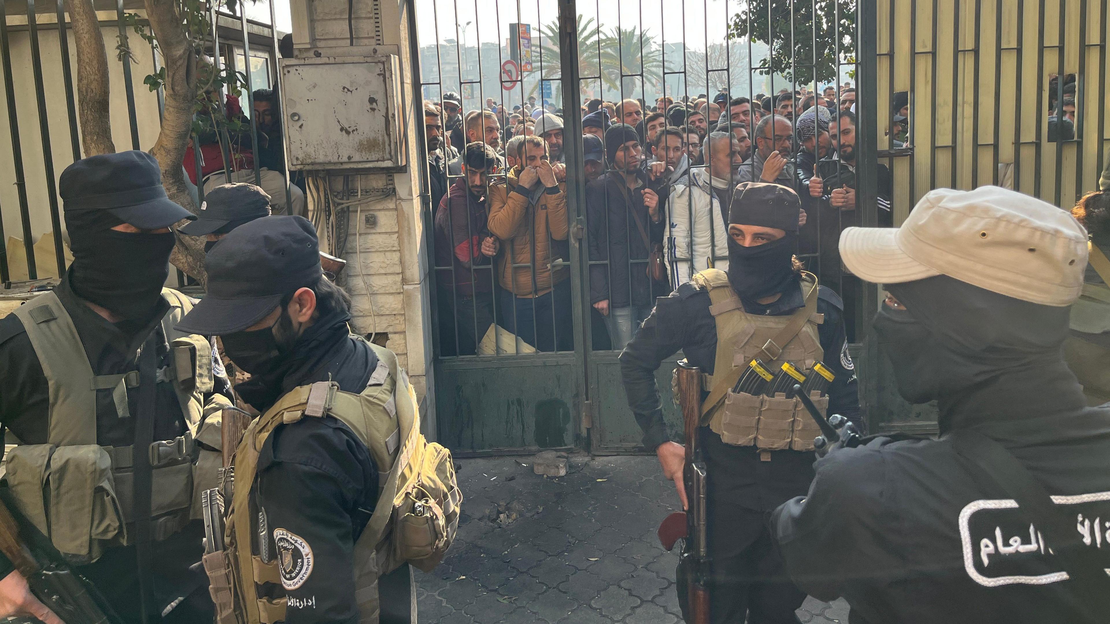 Armed and masked men stand inside a gate, while a crowd of other men press against the gate.