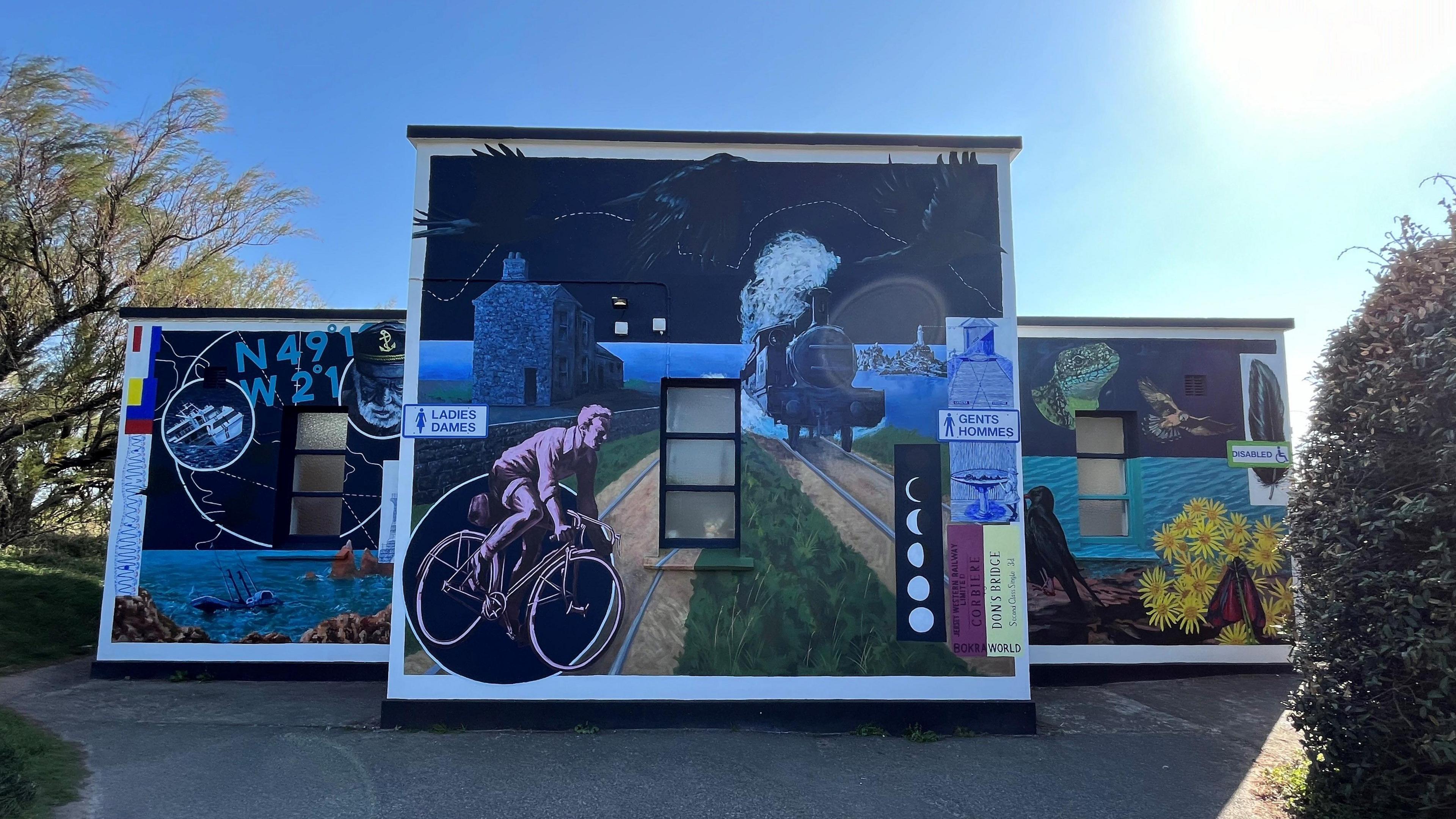 A three piece mural at a public toilet in Jersey - first square shows the ocean and other signs over it, second and main square shows a road with a cyclist to the left and a train on the right, the third shows the ocean and sunflowers. Sky behind is blue and clear.