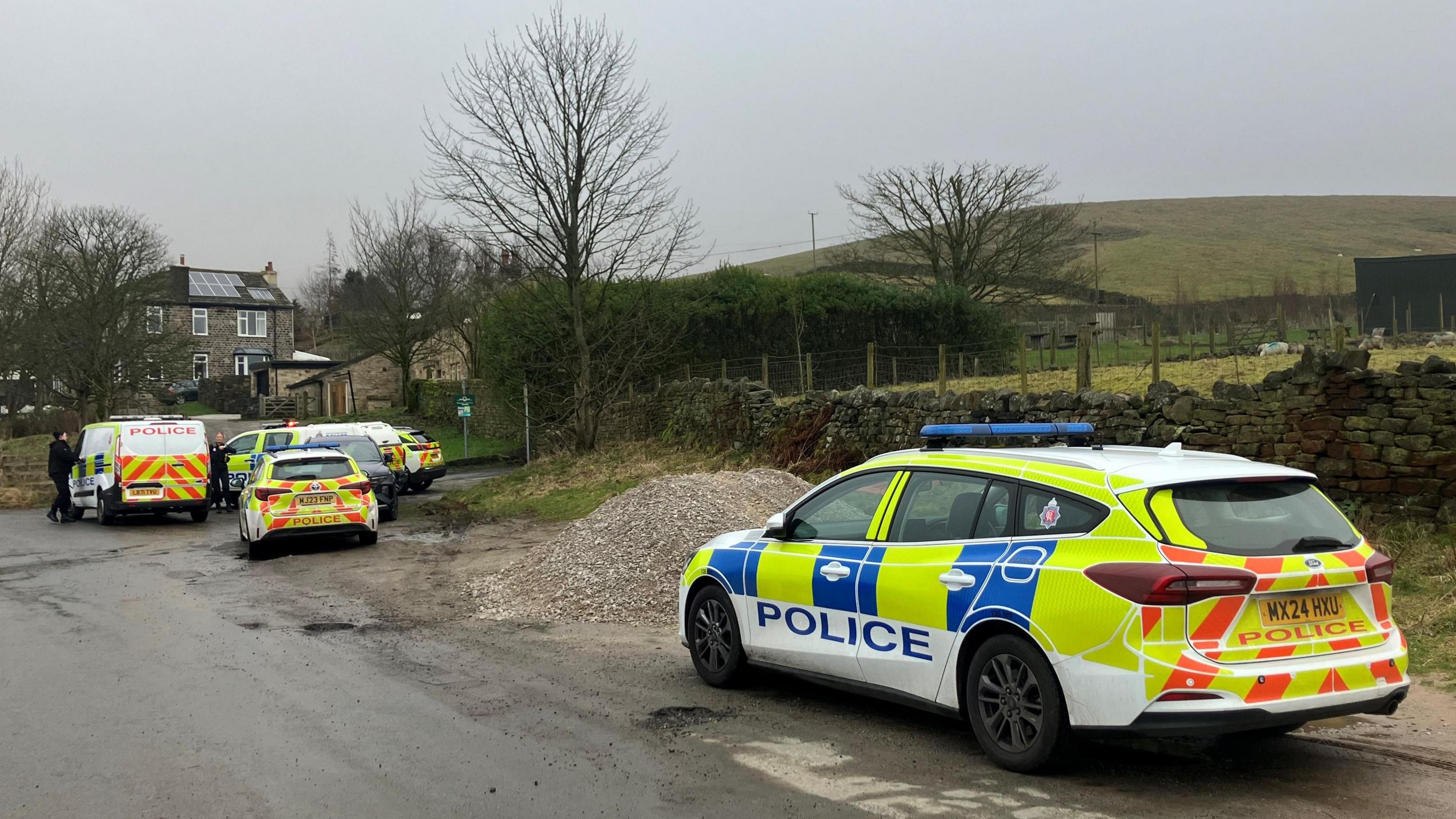 Police cars parked in a country lane n=clsoe to a dry stone wall, a field and trees that have shed their leave.