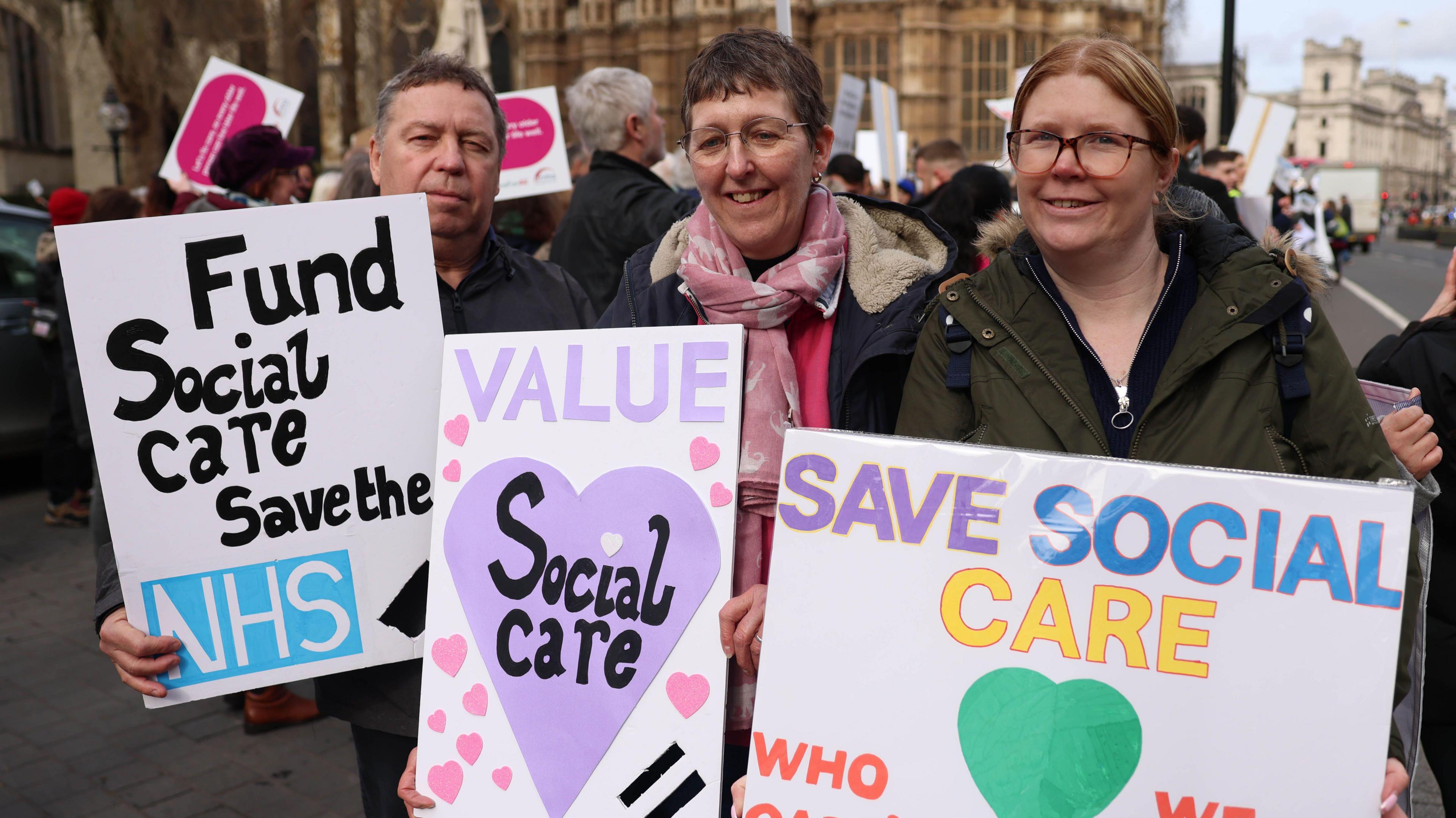 A man standing next to two women all holding placards calling for the government to save, value and fund social care