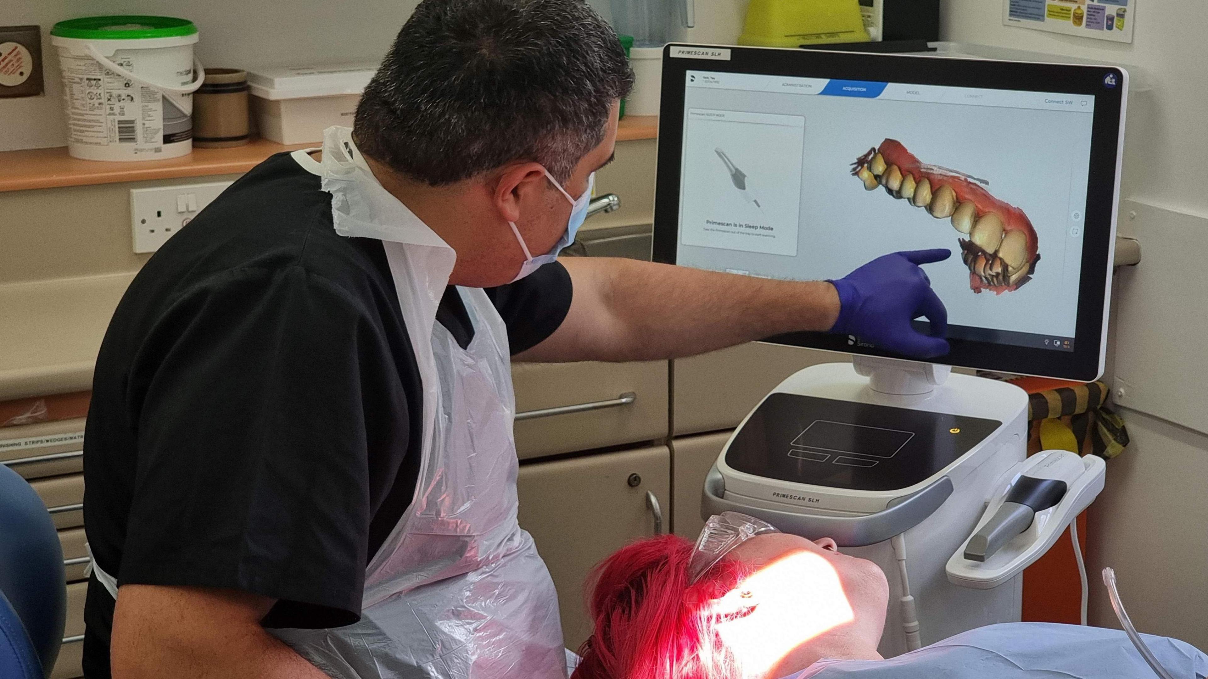 A consultant in black T-shirt pointing to a TV monitor with a 3-D colour image of a patient's gums and teeth on the screen.