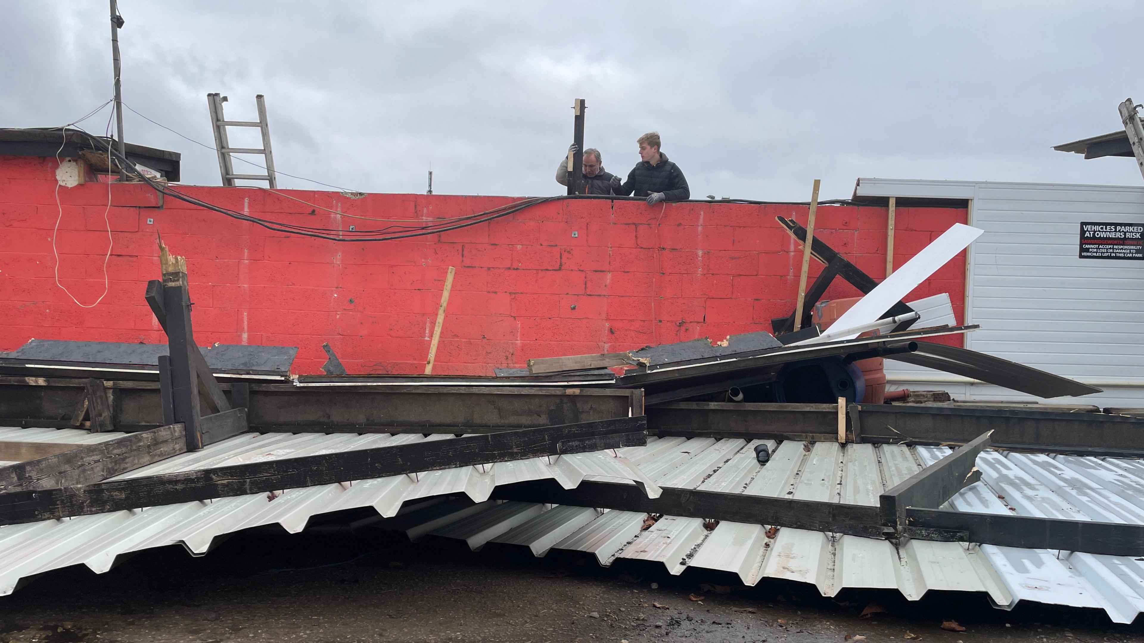 A red wall with a ladder propped against it. A sheet metal roof can be seen on the ground. Two men are looking over the damage.