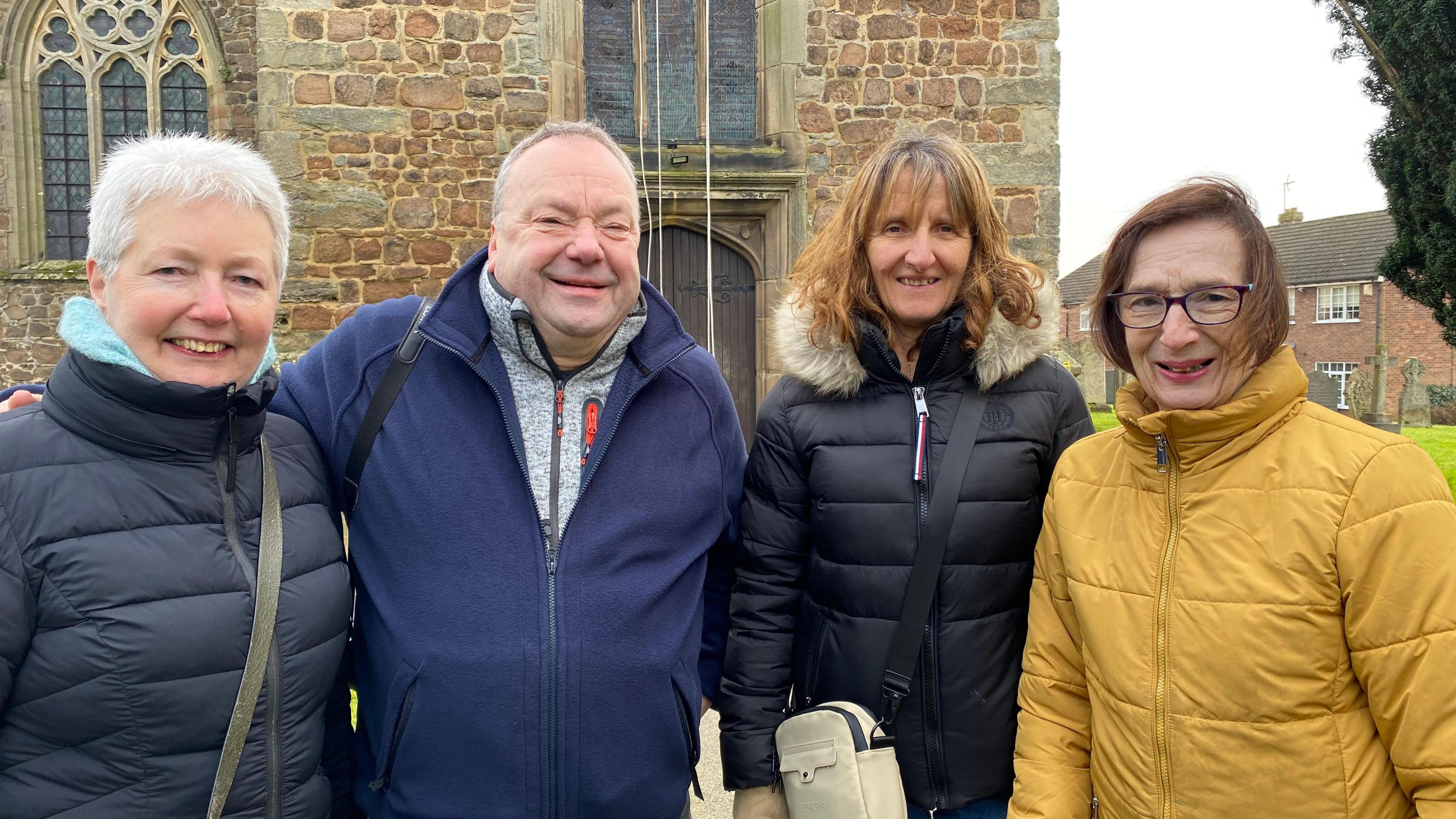 Three women and a man smile for the camera, with a oak coloured Church in the background. 