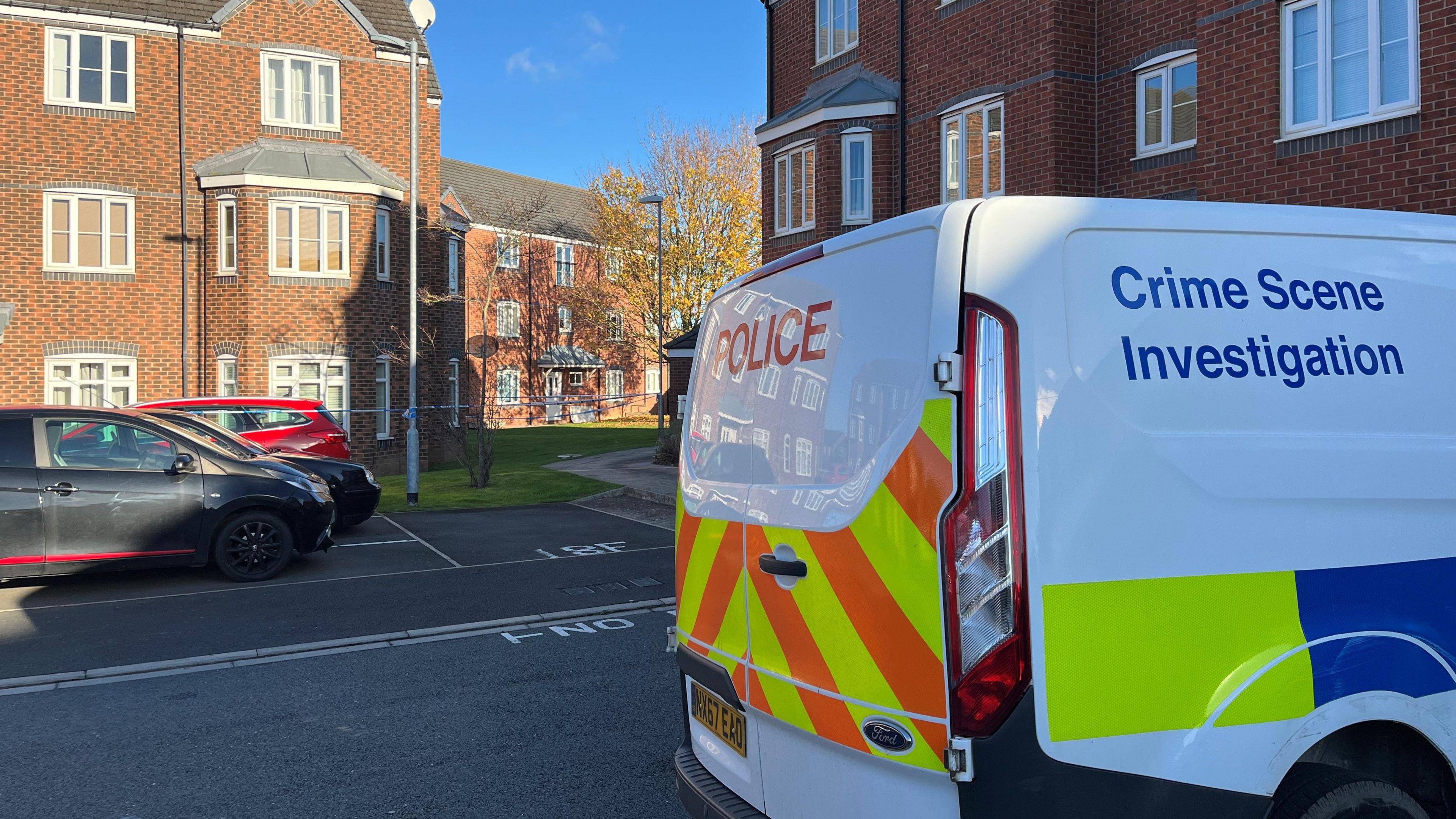 A police van with Crime Scene Investigation written on its side, stands parked on a housing estate, surrounded by large, red-brick buildings. Police tape can be seen in the distance and there are parked cars to the left of the van.