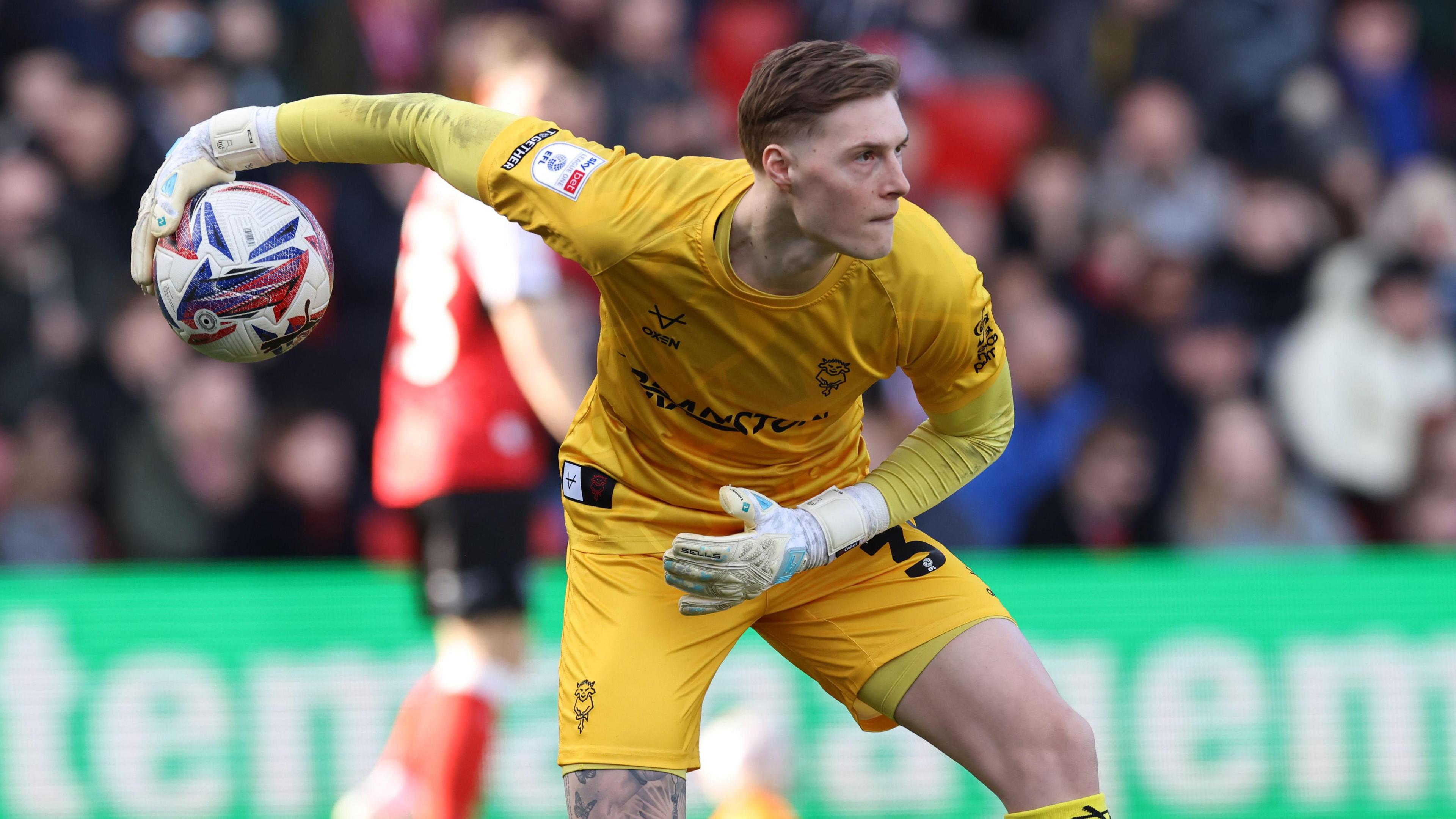 Zach Jeacock is all in yellow and preparing to roll the ball out to a team-mate during a match for Lincoln City. There is an opposition player in orange behind him against the dark background of the crowd.