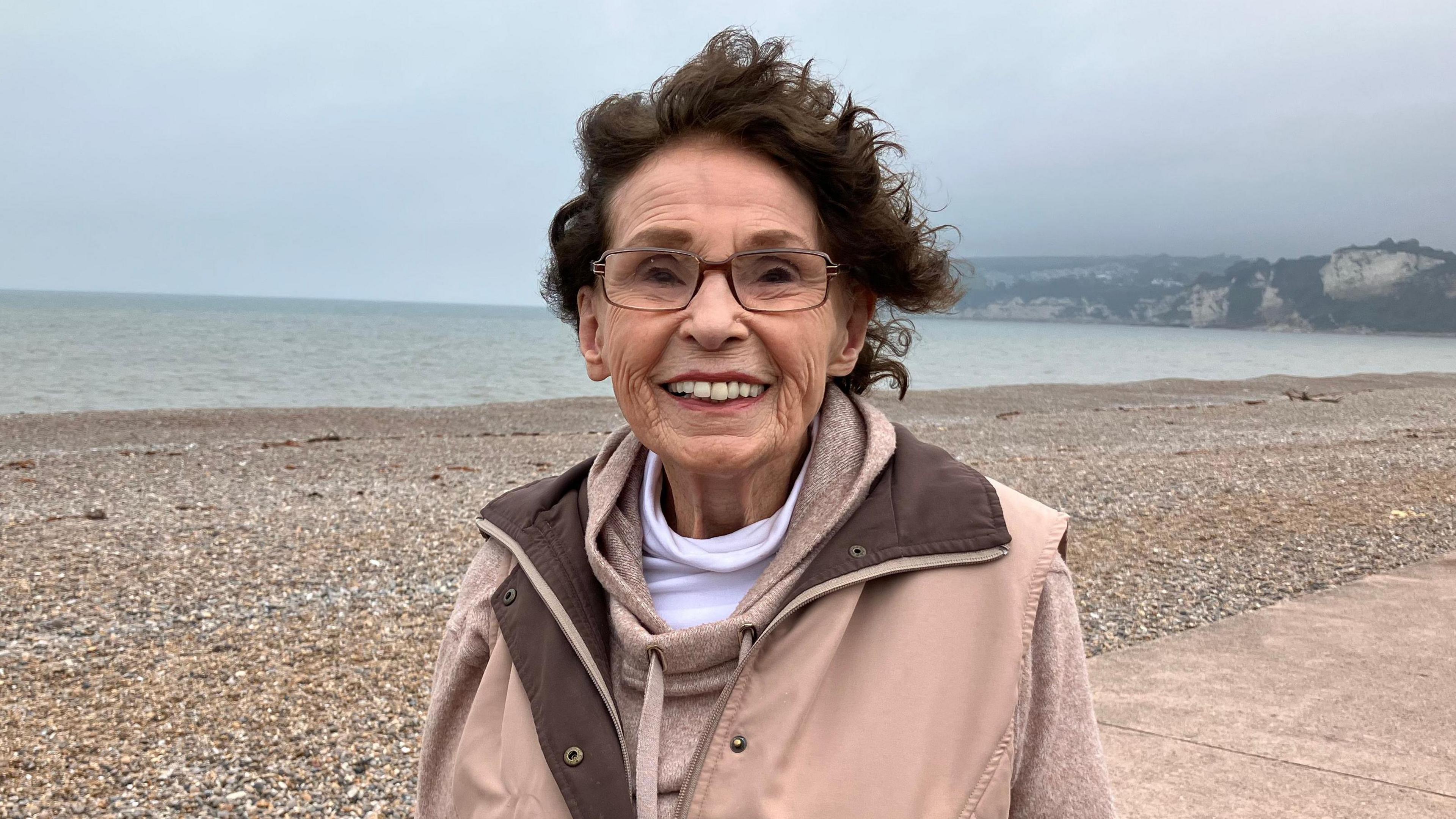 Joan Norcombe smiling while walking along Seaton seafront on a cloudy day. She is stood on an esplanade in front of a stony beach.