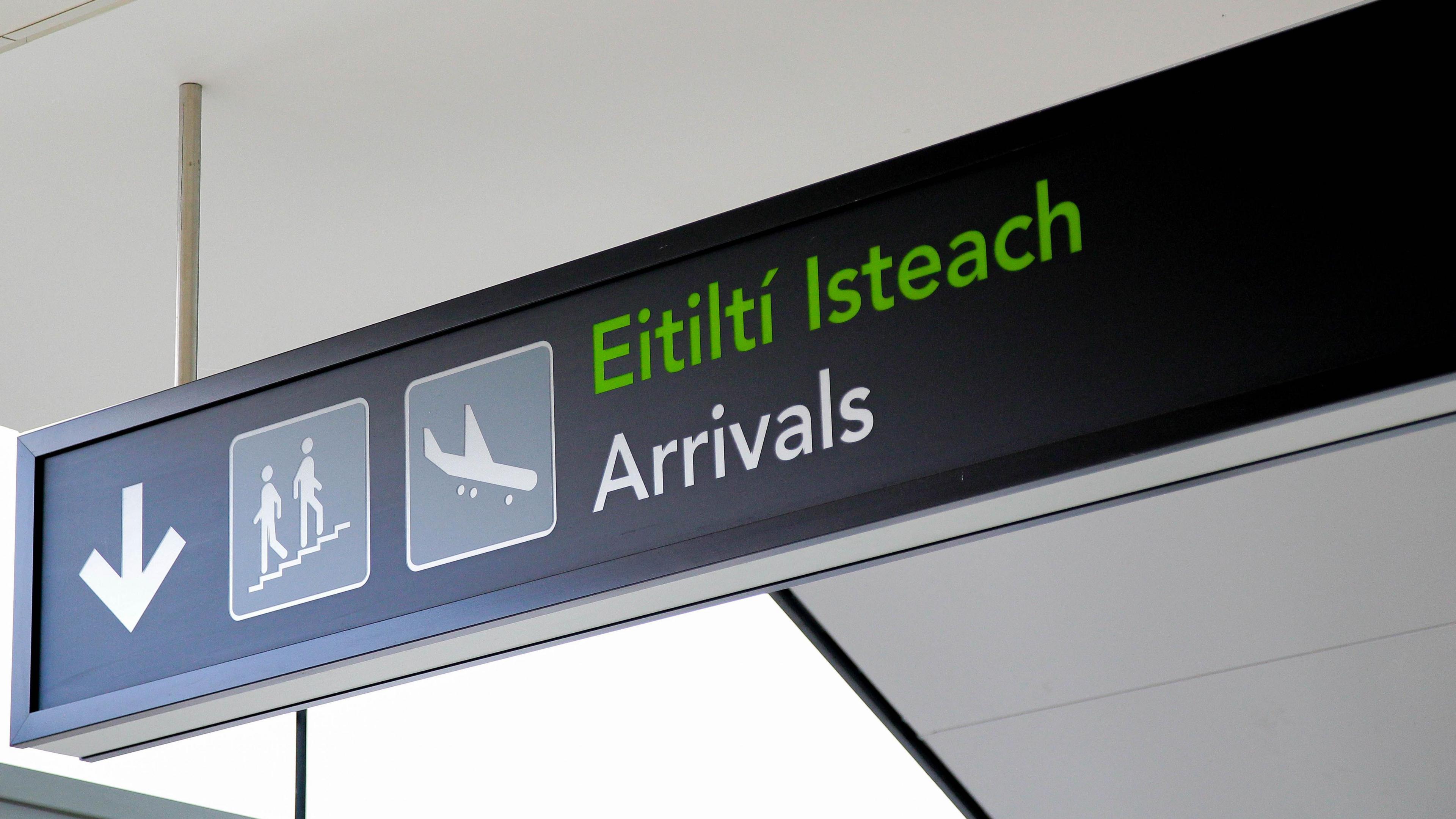 A black sign reads "arrivals" with the Irish translation written above in green. Beside the text is an image of a plane, and people descending a staircase beside a white arrow pointing downwards
