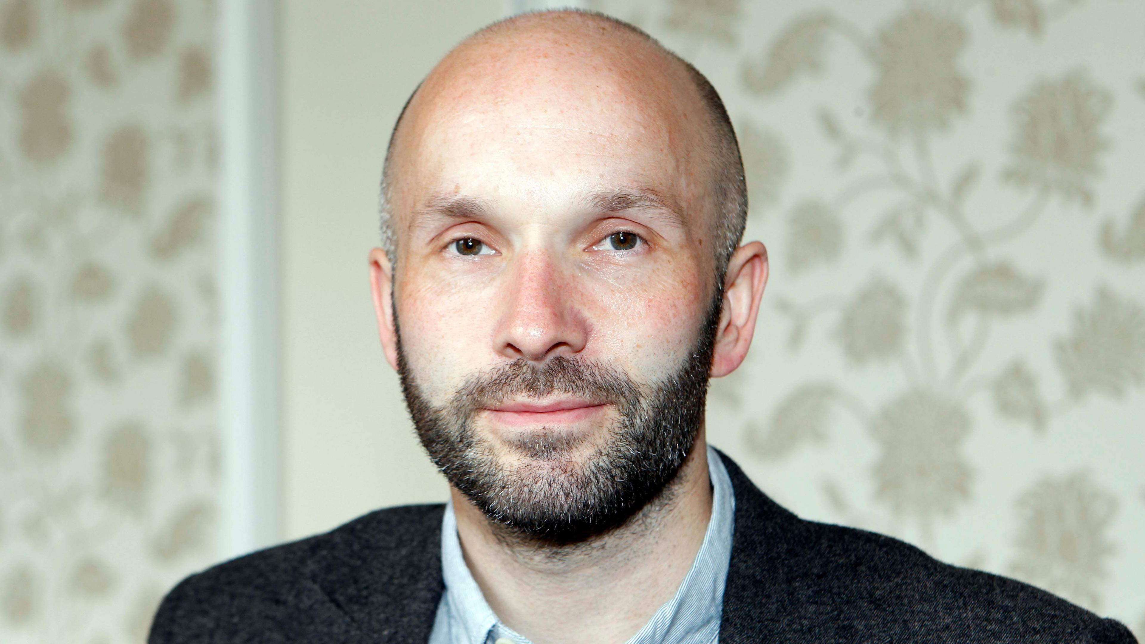 A man in his forties with a shaved head and a short beard. Flowery wallpaper is behind him and he is wearing a suit.