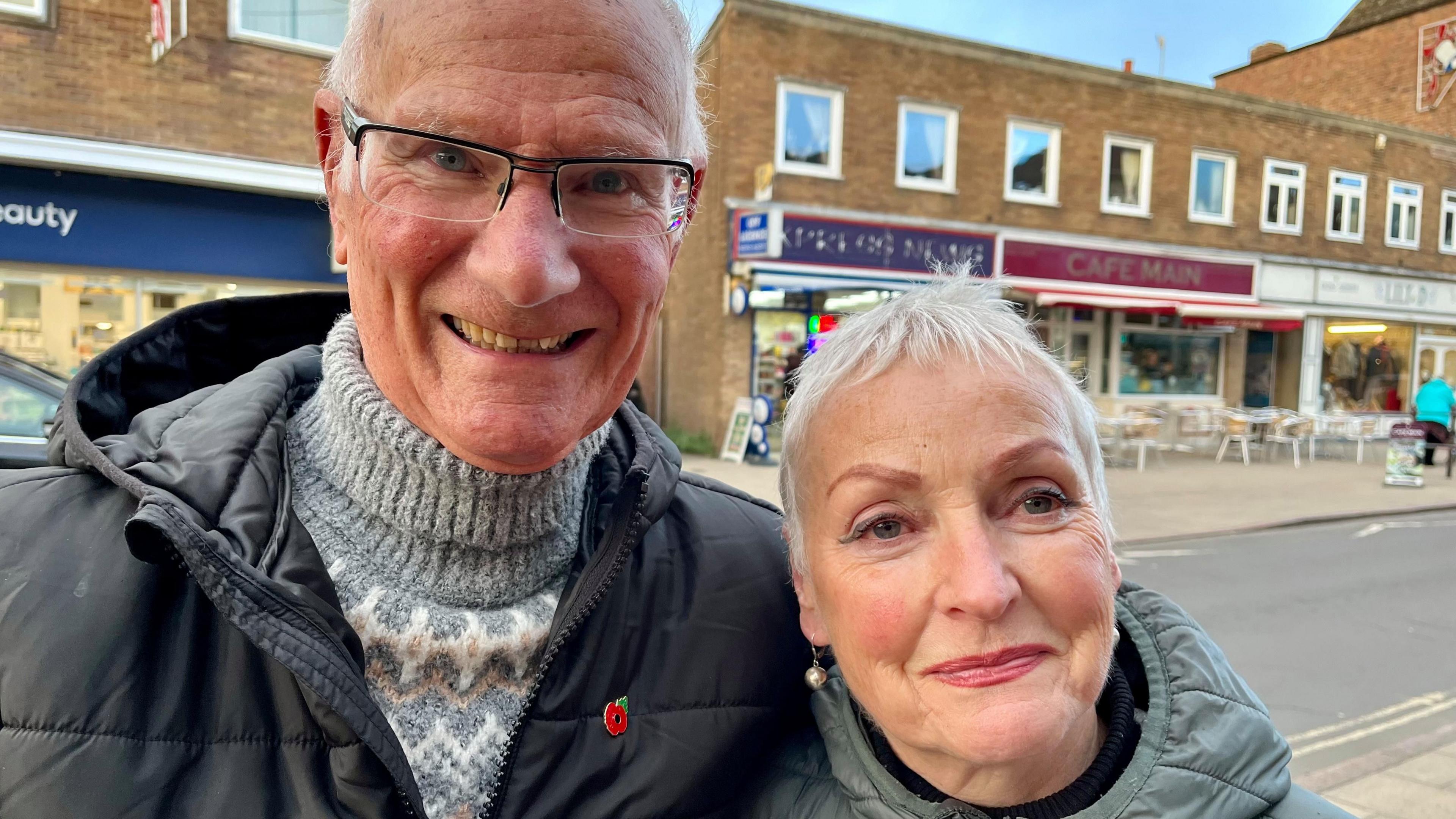 Gill Ormesher (right) is standing in Cromer town centre with shops behind her. She is wearing a grey hooded coat and is standing next to her husband who has a black coat, grey jumper and is wearing a poppy pin badge.