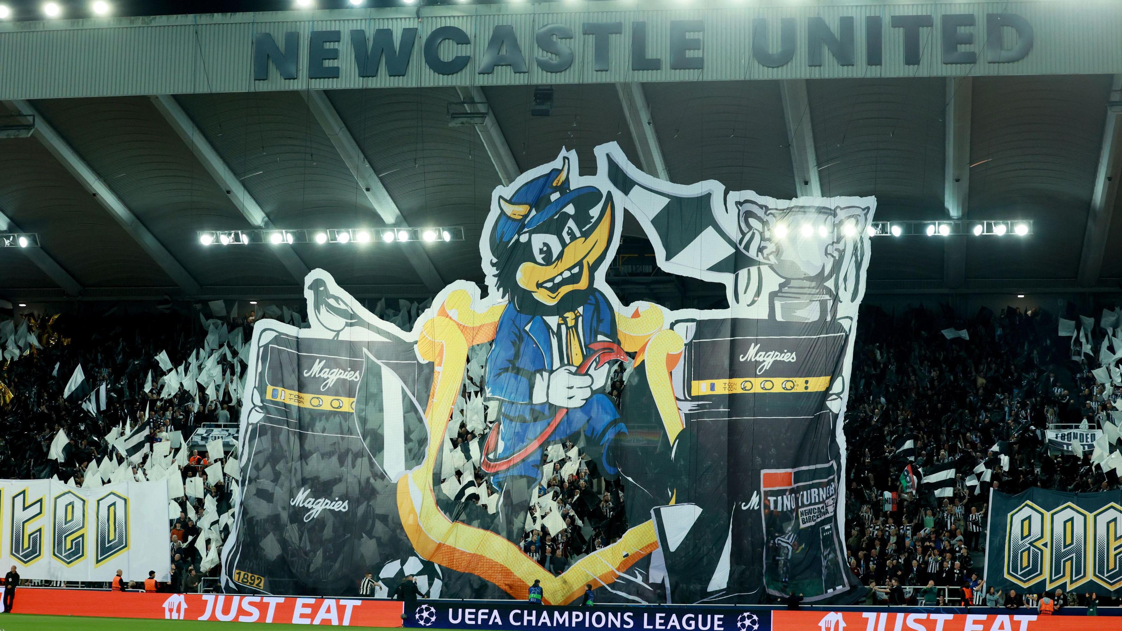 A giant flag of a tifo (a blue and black bird-like mascot) standing in front of speakers based on the Marshall design but with the company's name replaced with the word Magpies. The flag spans the height of St James' Park football stadium. Stands-full of fans hold black and white flags behind the display.