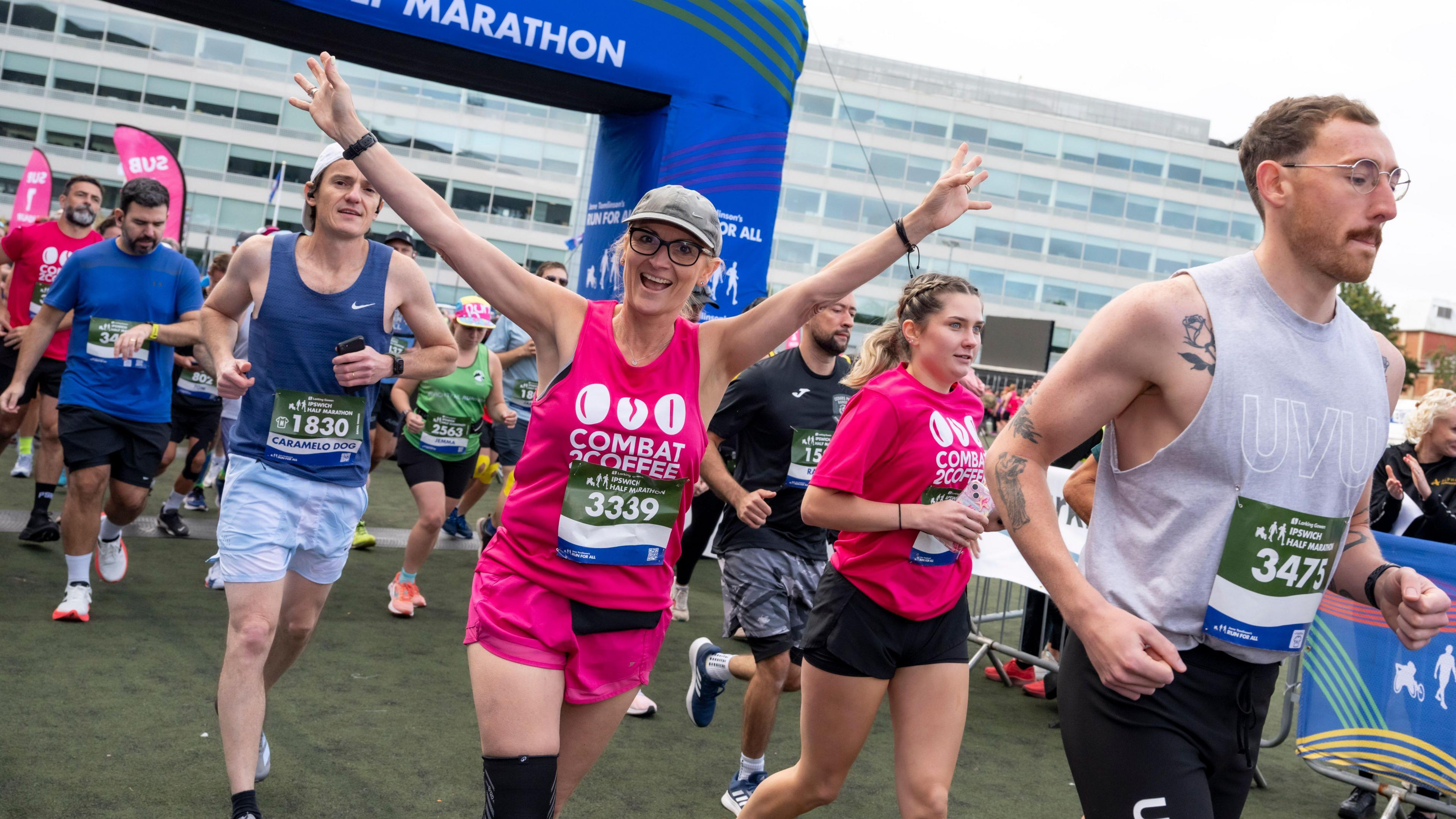 A woman dressed in pink is running a marathon while cheering, her arms in the he air, others around her focused on the run ahead