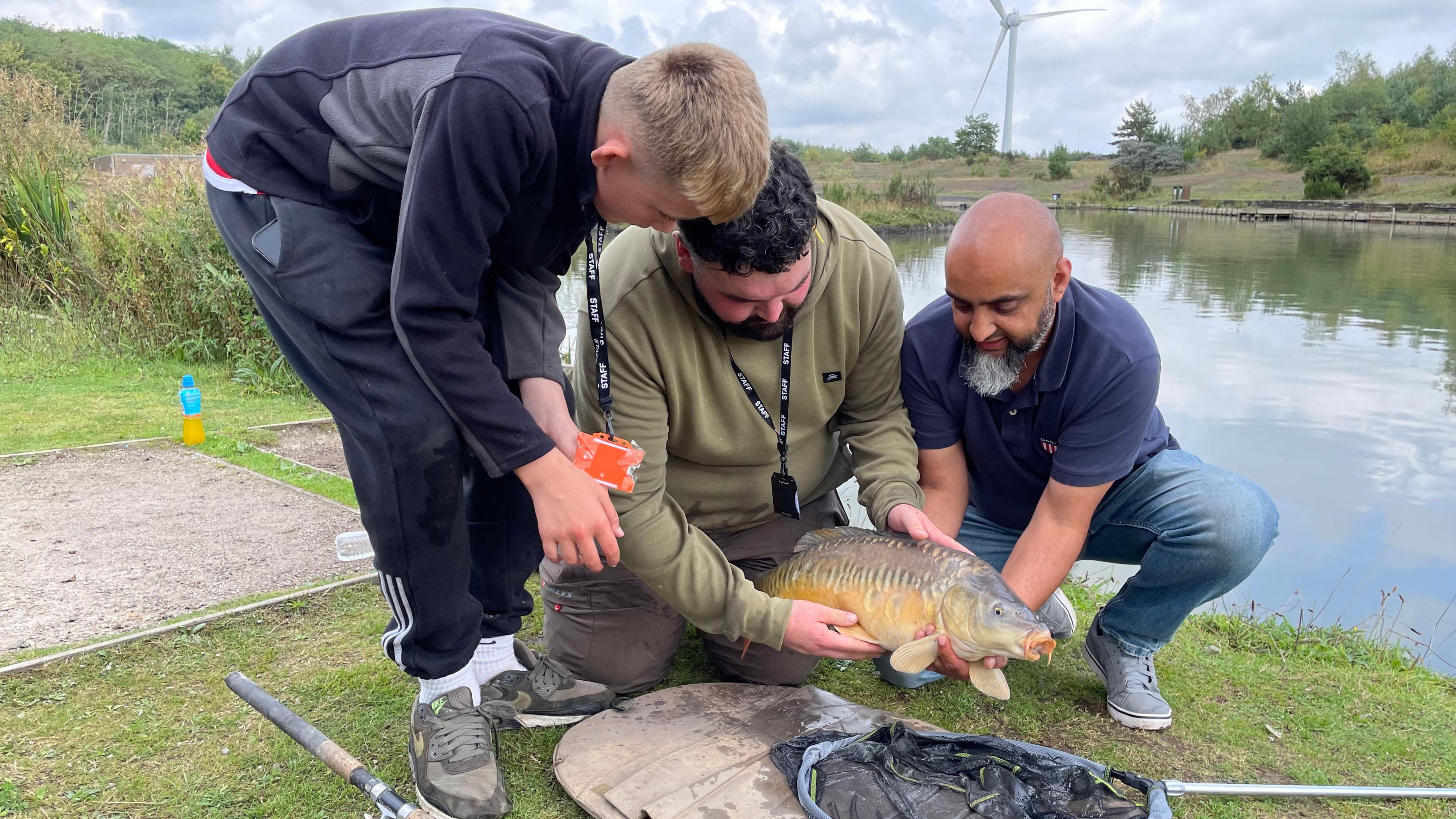 Three males - one stood up and bent over, one kneeling and another crouch down - hold a freshly-caught fish at the angling lake near Newstead. A wind turbine is in the background beyond the edge of the water