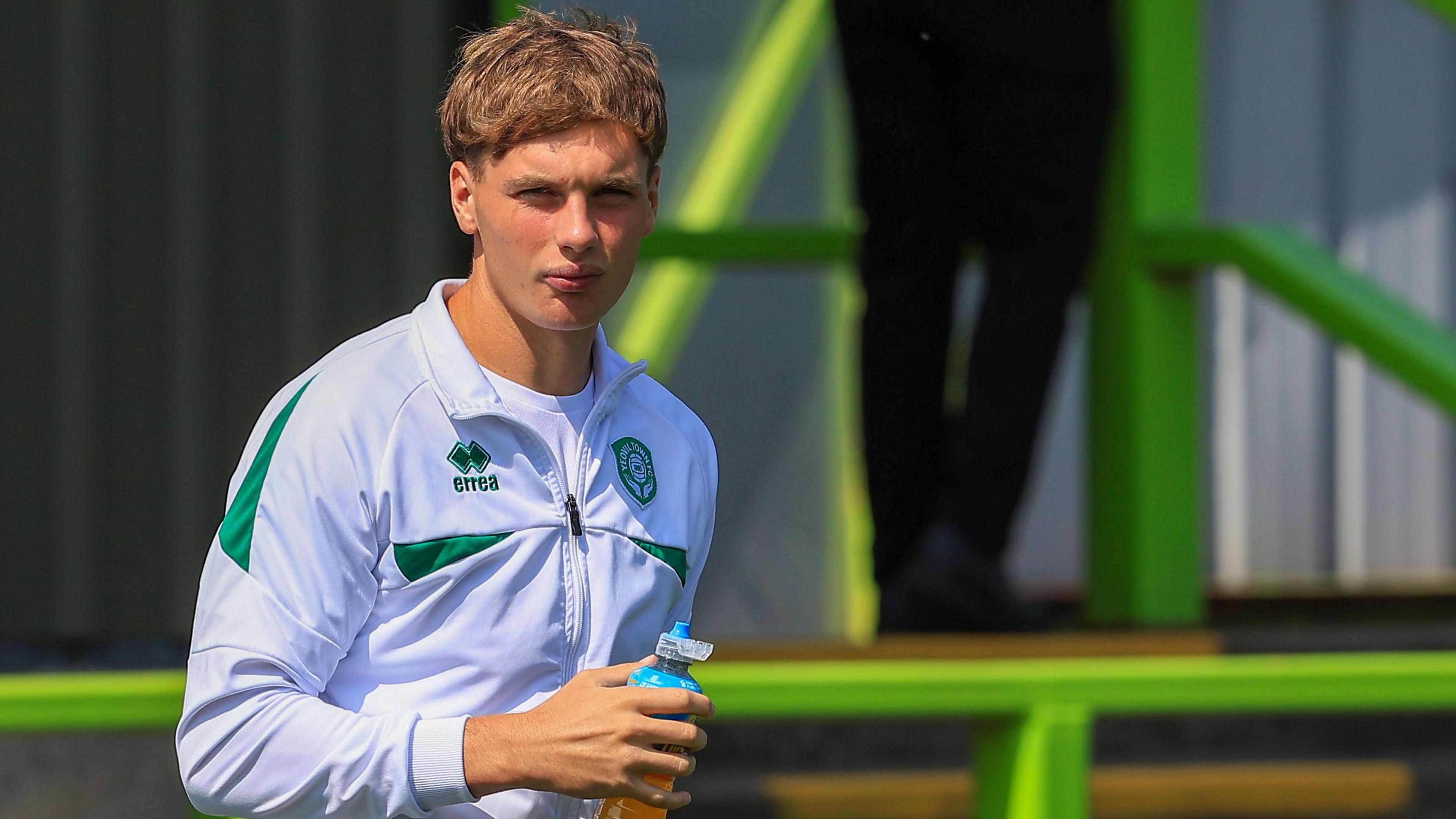 Ben Wodskou watches on during a break in a pre-match warm-up for Yeovil Town. He is wearing the club training zipper and is holding a sports drink in his right hand. 