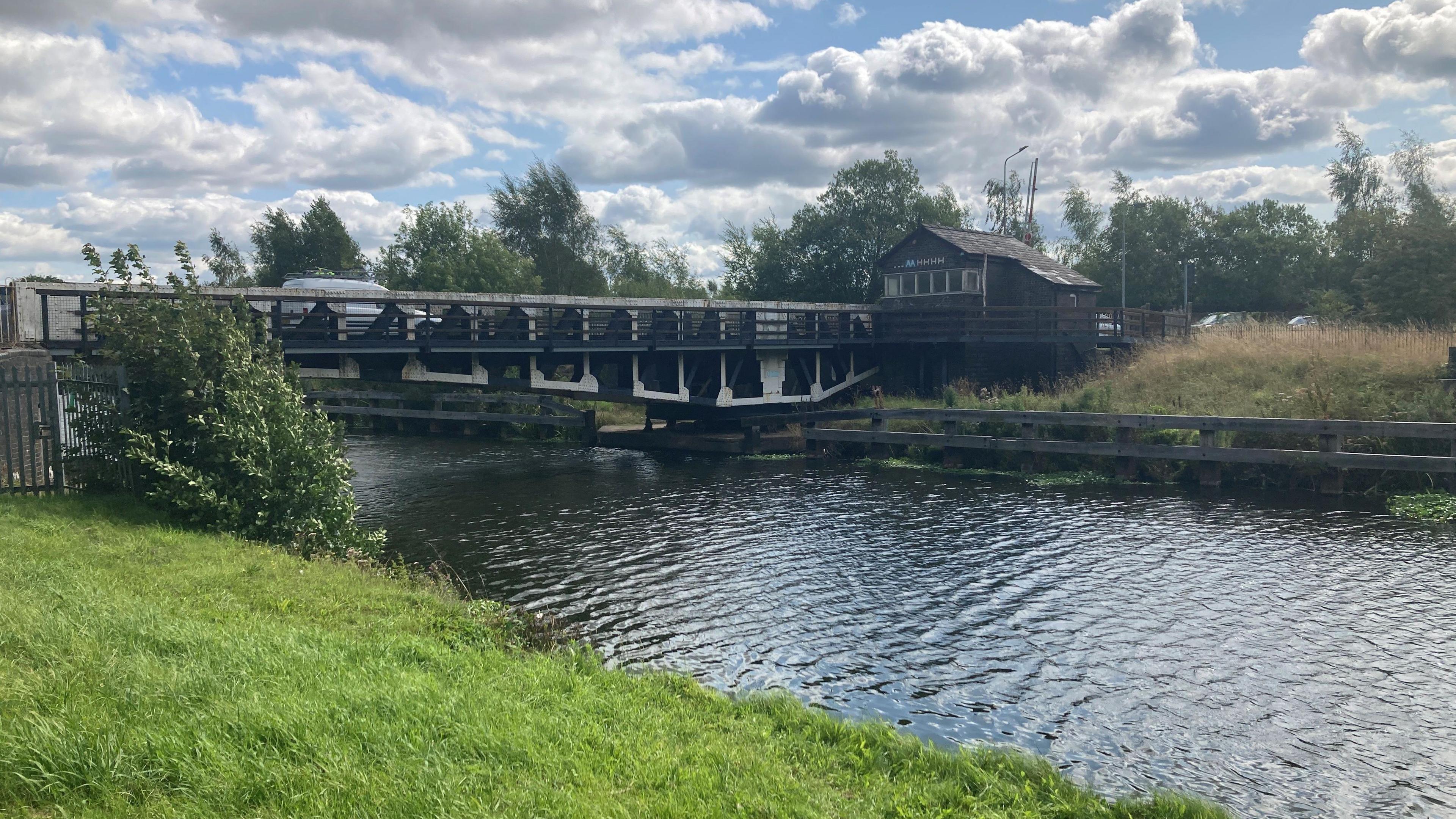 A view of the Winnington Swing Bridge over the River Weaver. It is a short, metal bridge with a wooden hut at the end of it on the far bank. 