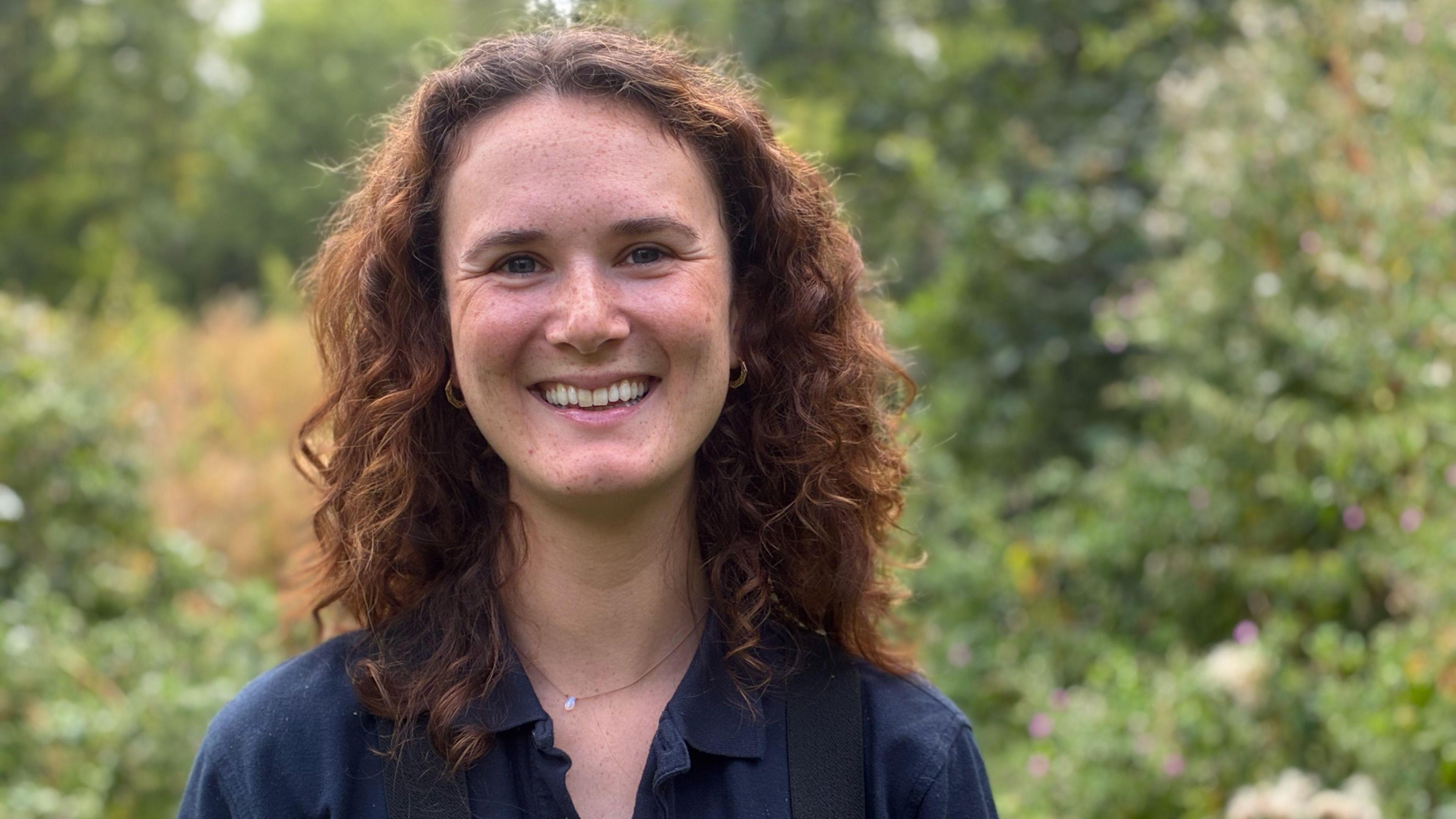 A woman with long curly red/brown hair smiles at the camera. She has a blue collared shirt on and there is a lot of green vegetation in the background.