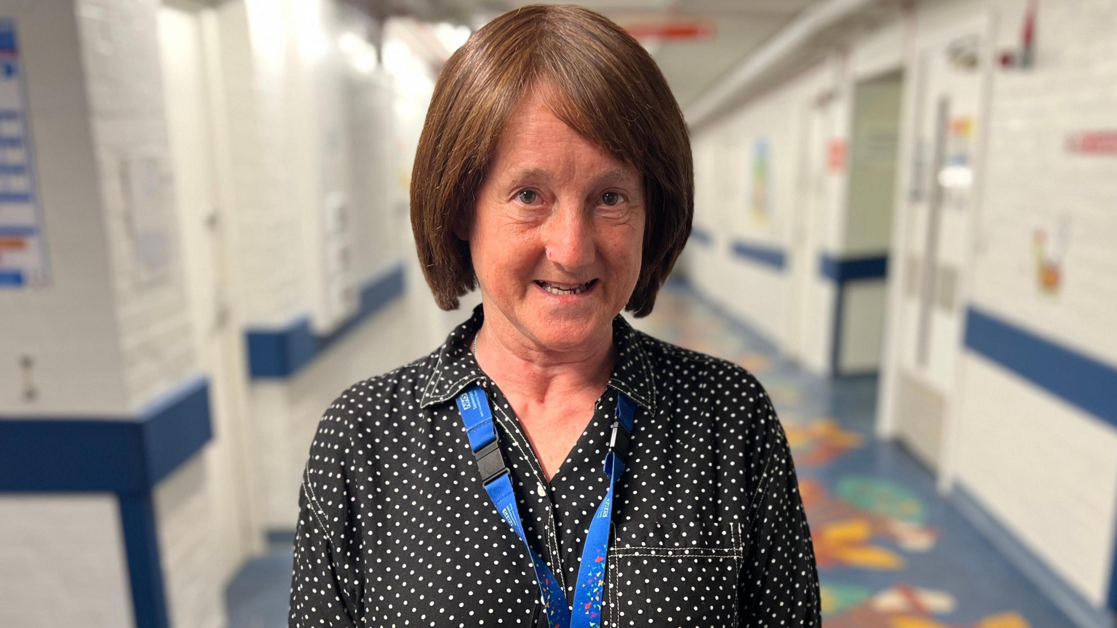 Helen Parfitt ears a black polka dot shirt and a blue lanyard in a hospital corridor. 