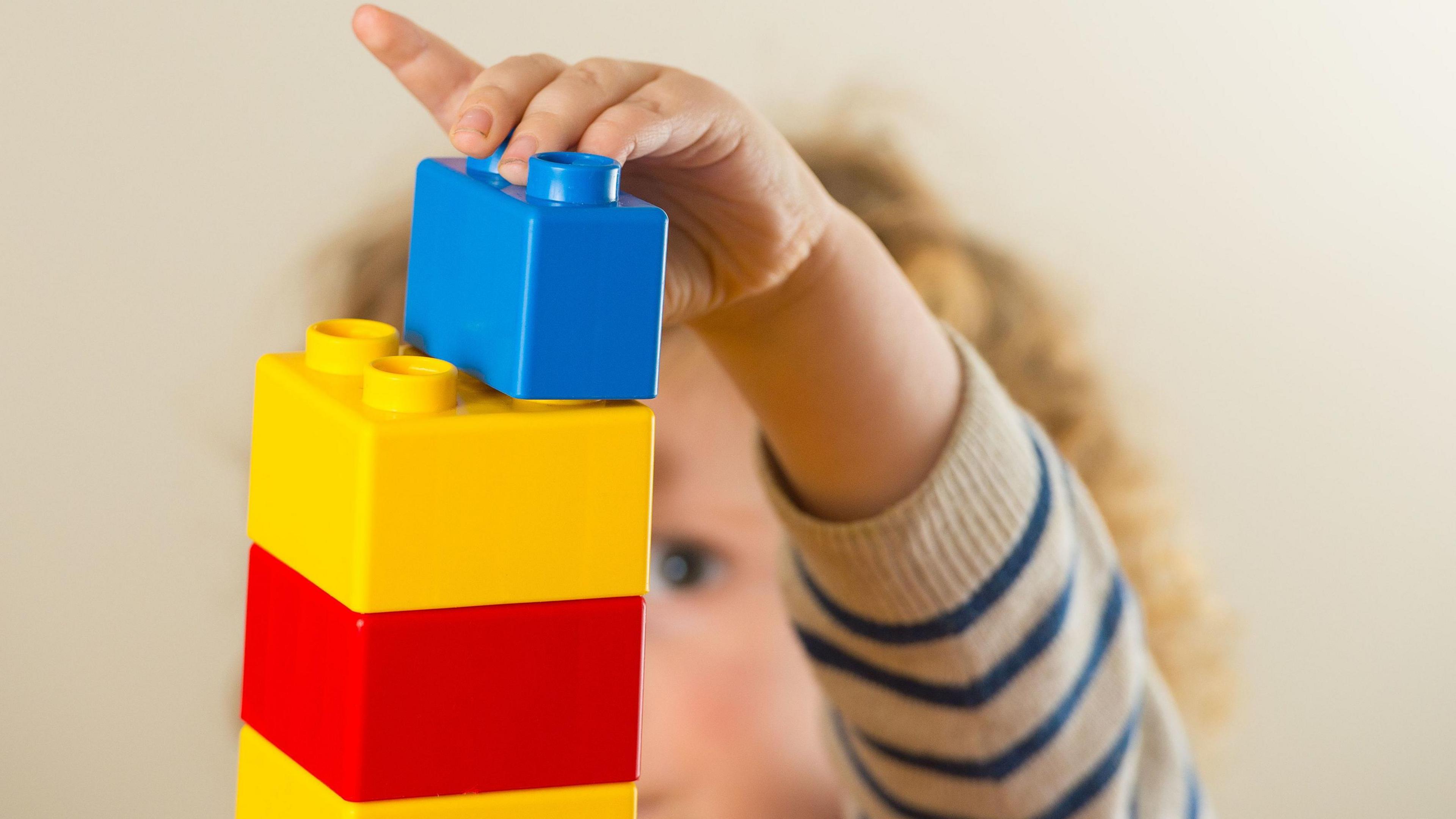 A toddler is building a structure from plastic building blocks. There is a small tower of yellow and red blocks with a blue one on the top.