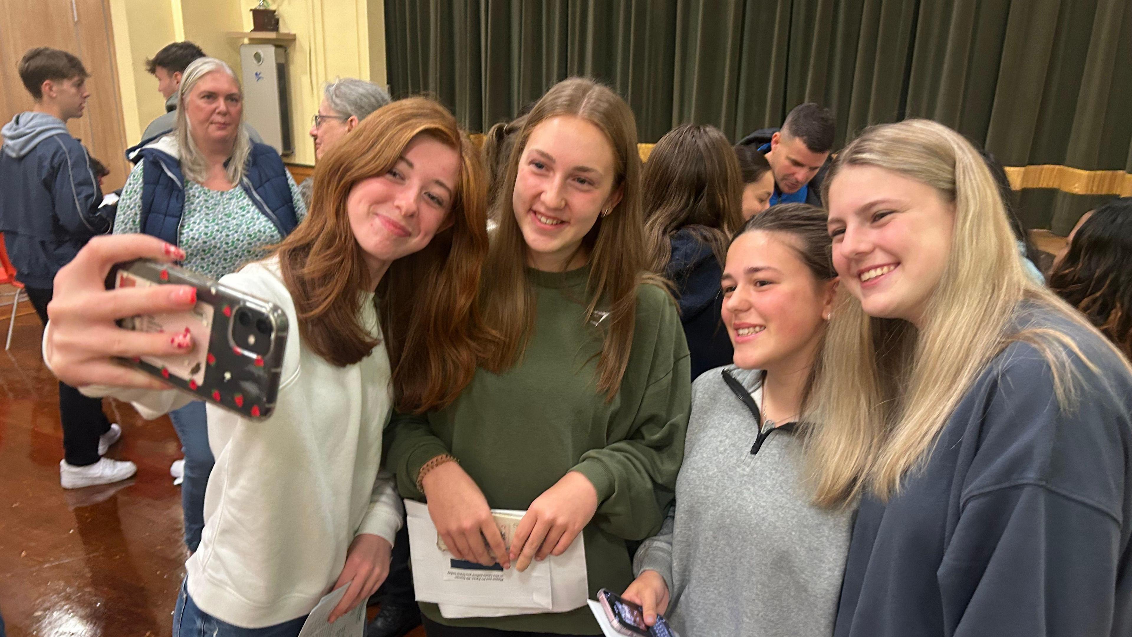 Four 16-year-old girls  smiling for a selfie in a school hall with dark green curtains in the background and other students.