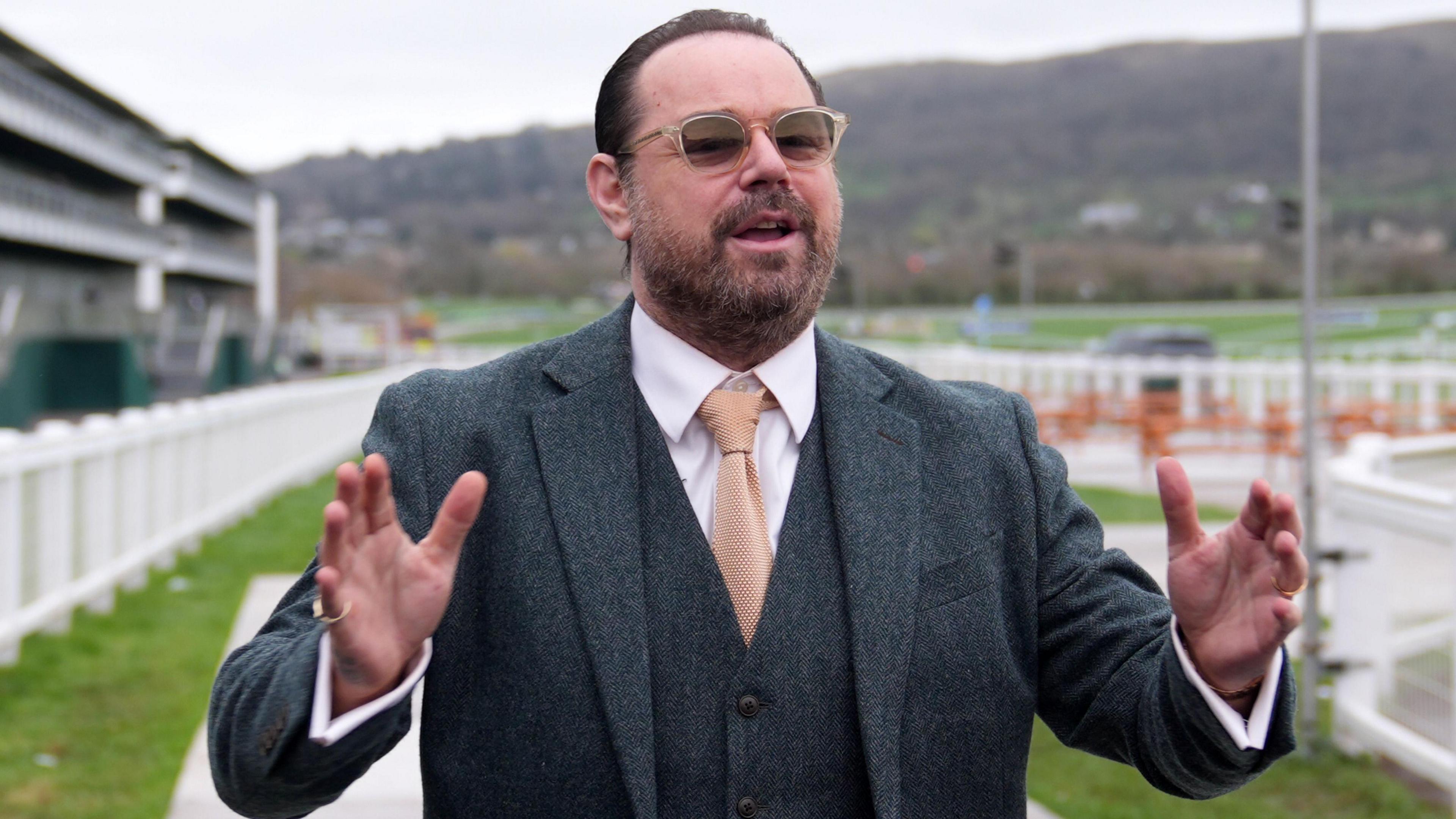 Actor Danny Dyer, wearing a dark grey jacket and waistcoat with a white shirt and light orange tie, holds his arms wide as he poses for photographers at Cheltenham Festival 2026