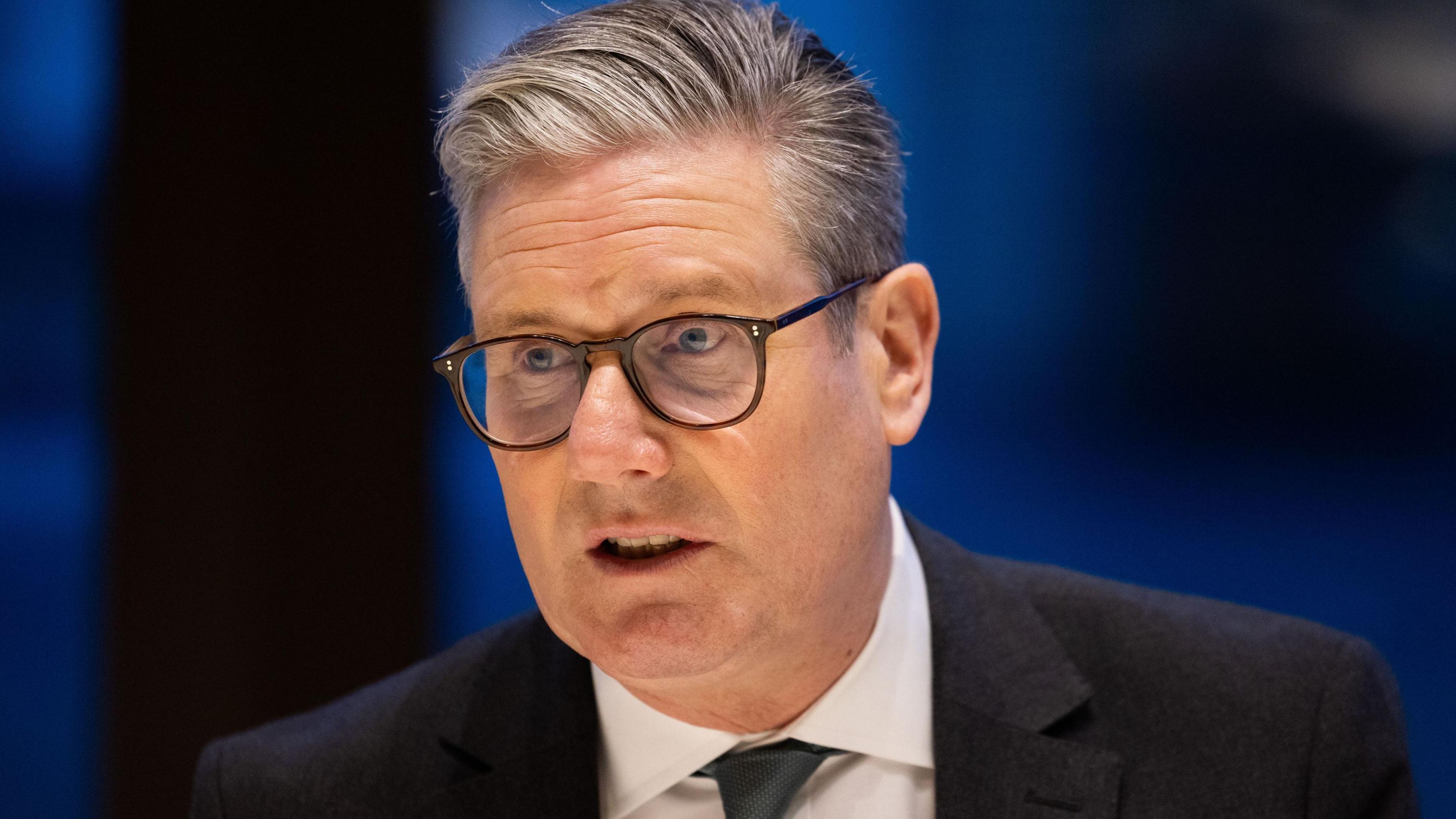 Sir Keir Starmer, in jacket and tie, and wearing glasses, in front of a dark-blue background