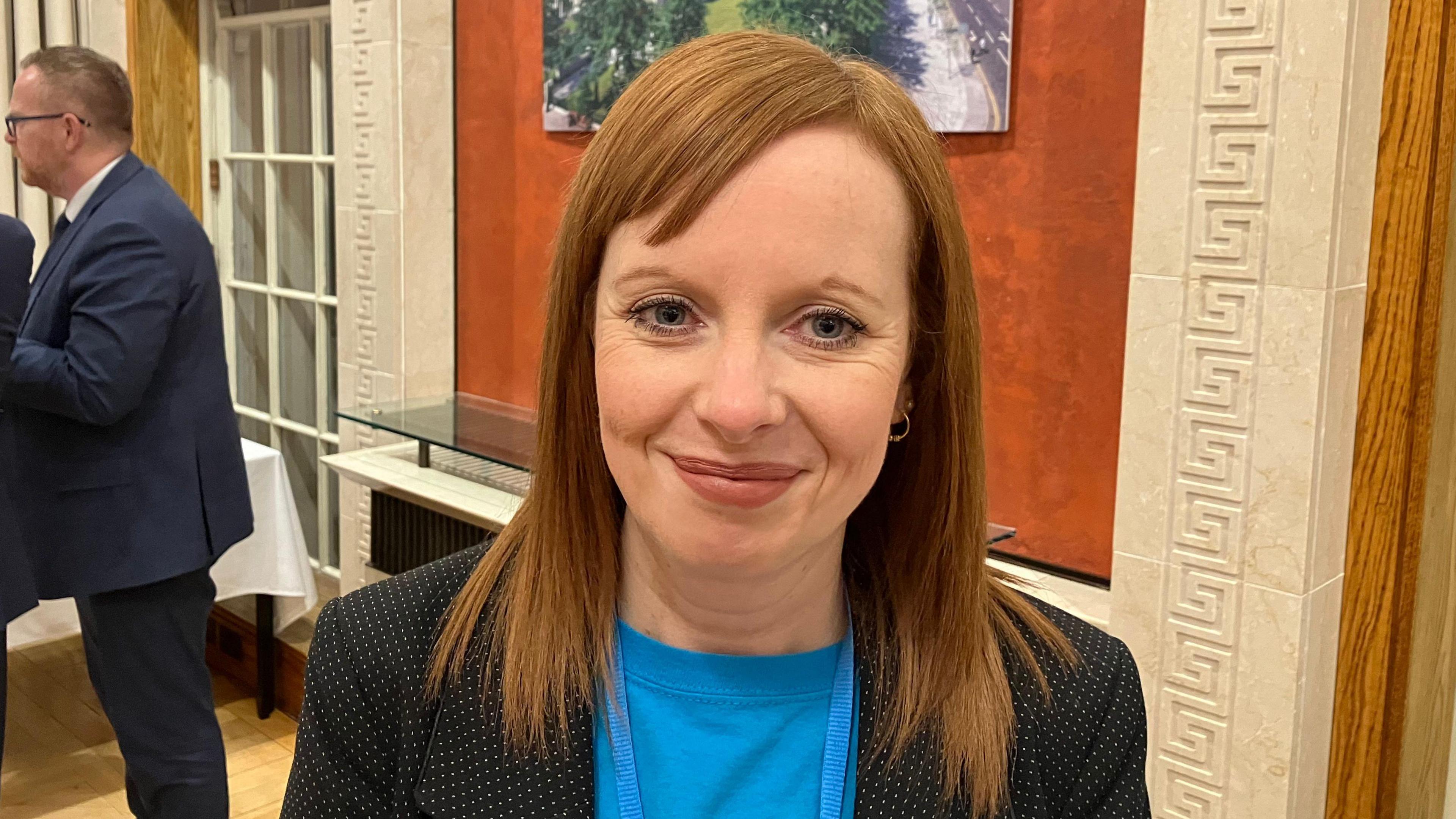 Alma White - a woman with shoulder-length, auburn hair looks at the camera. She is wearing a blue t-shirt and a black and white polka dot suit jacket.