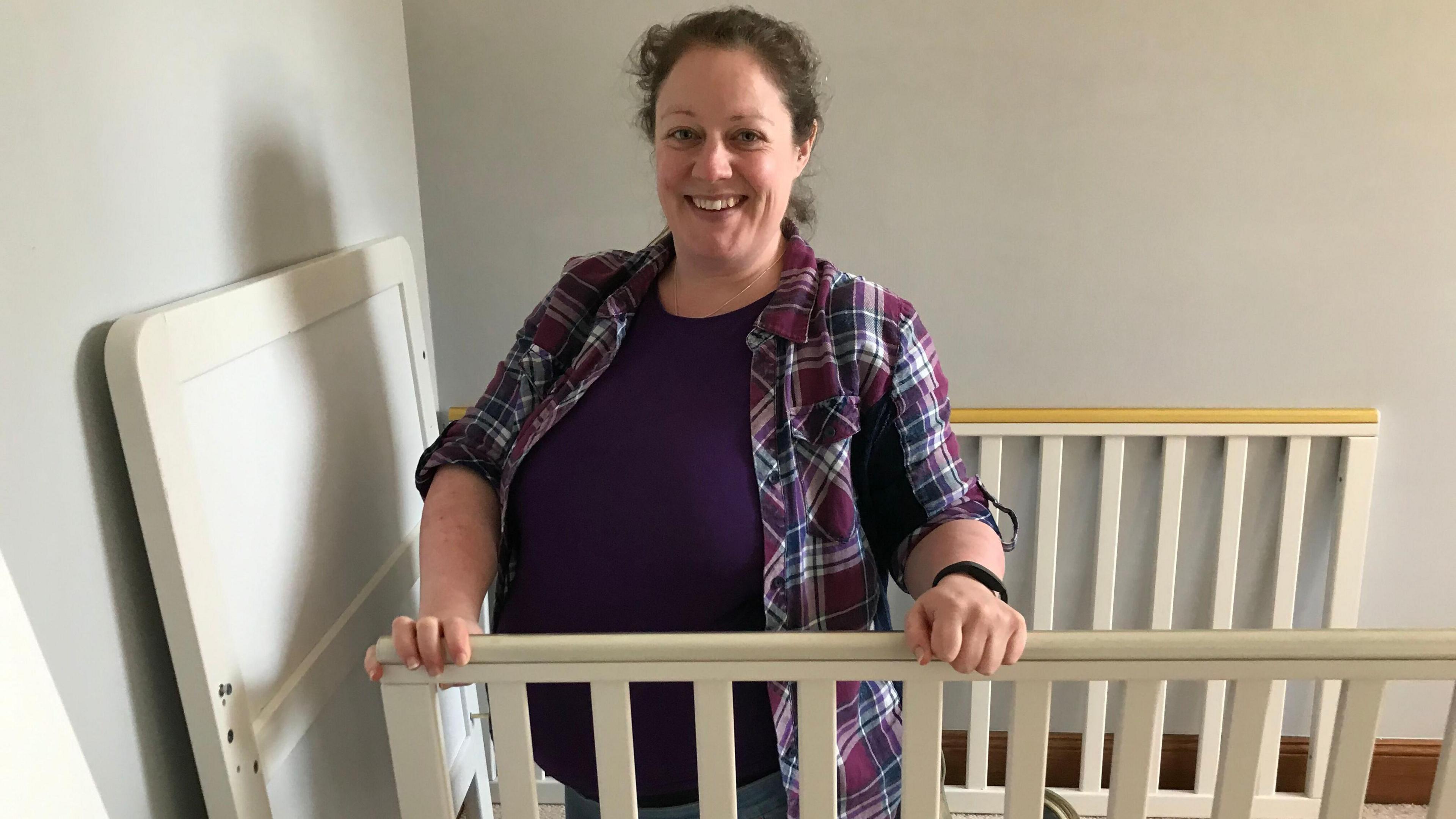 Jacqui Hunter, who is heavily pregnant. She is wearing a checked shirt and smiling at the camera is holding the sides of a baby cot that is being assembled