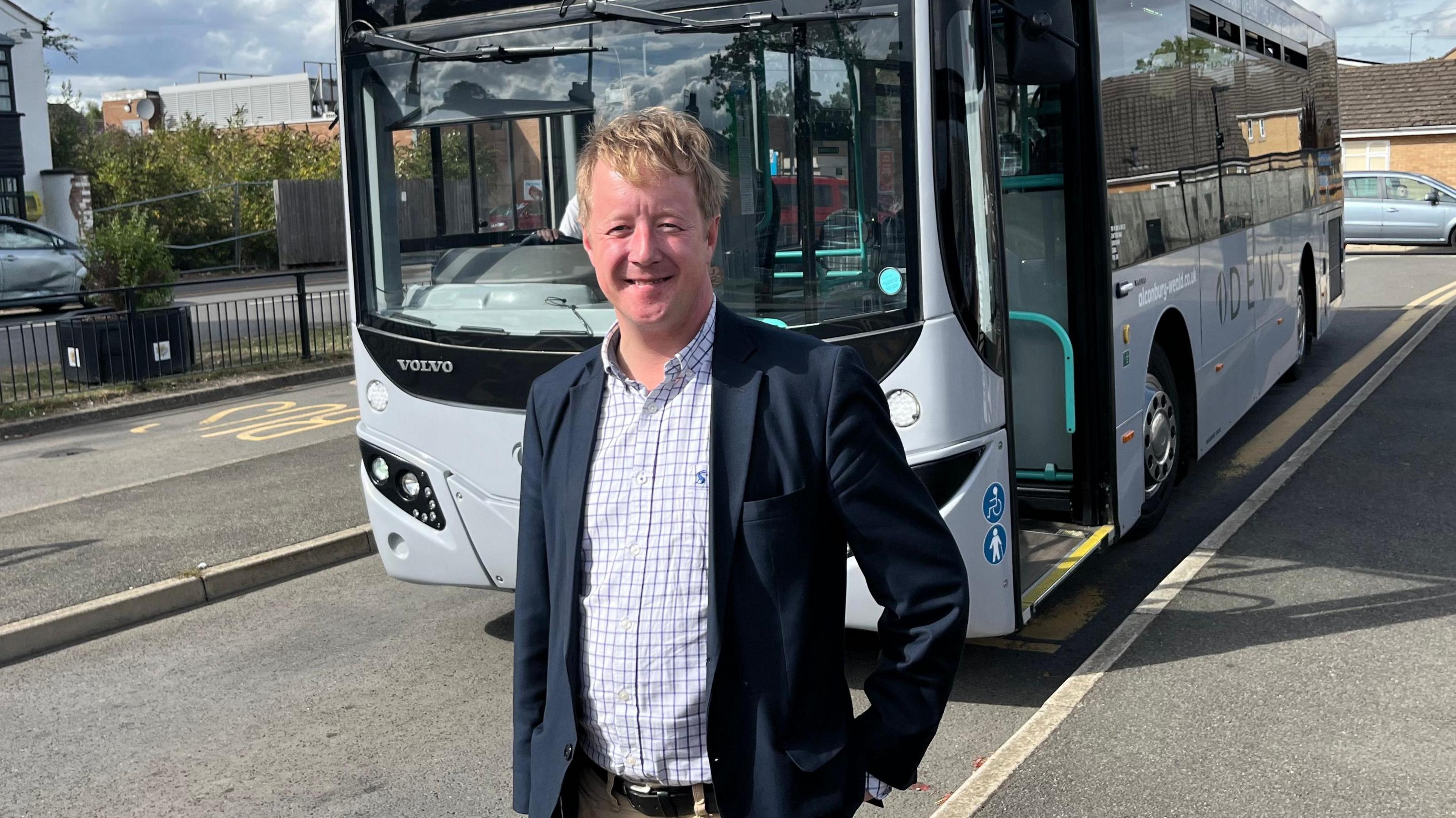 Paul Bristow, who is wearing a navy suit and white checked shirt, smiling at the camera and standing in front of a stationary grey bus which is on a road.