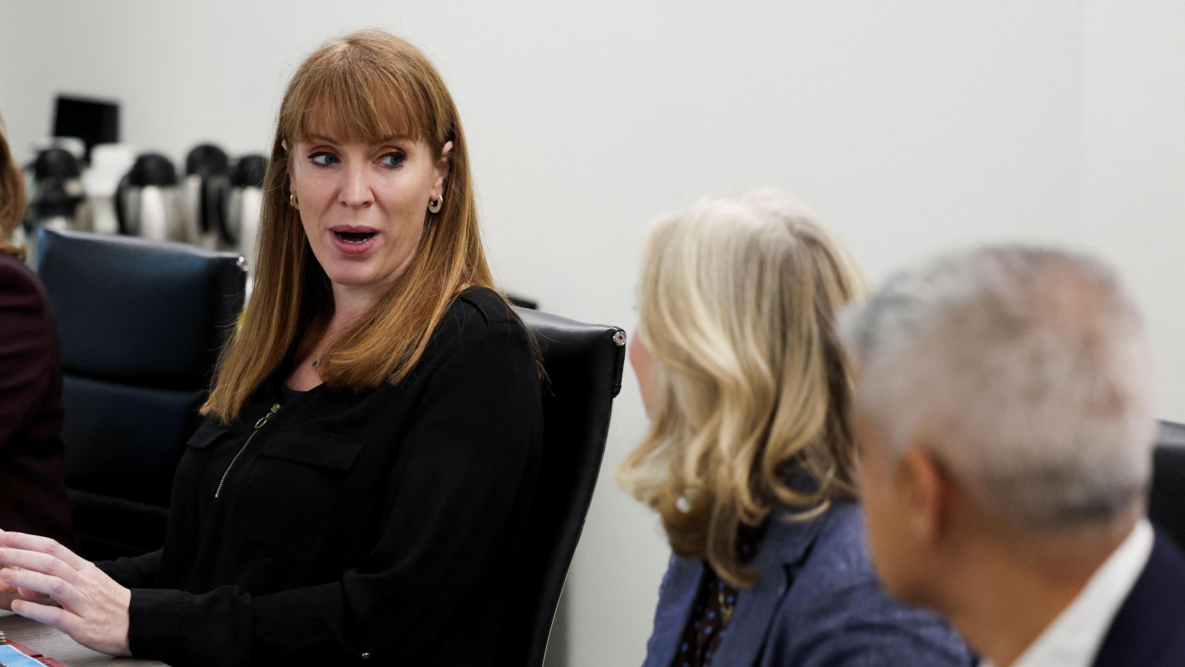 Deputy prime minister Angela Rayner chairs a meeting of elected mayors. She sits in the middle of a board room table addressing the other attendees. She wears a black suit and has long ginger hair with a fringe.