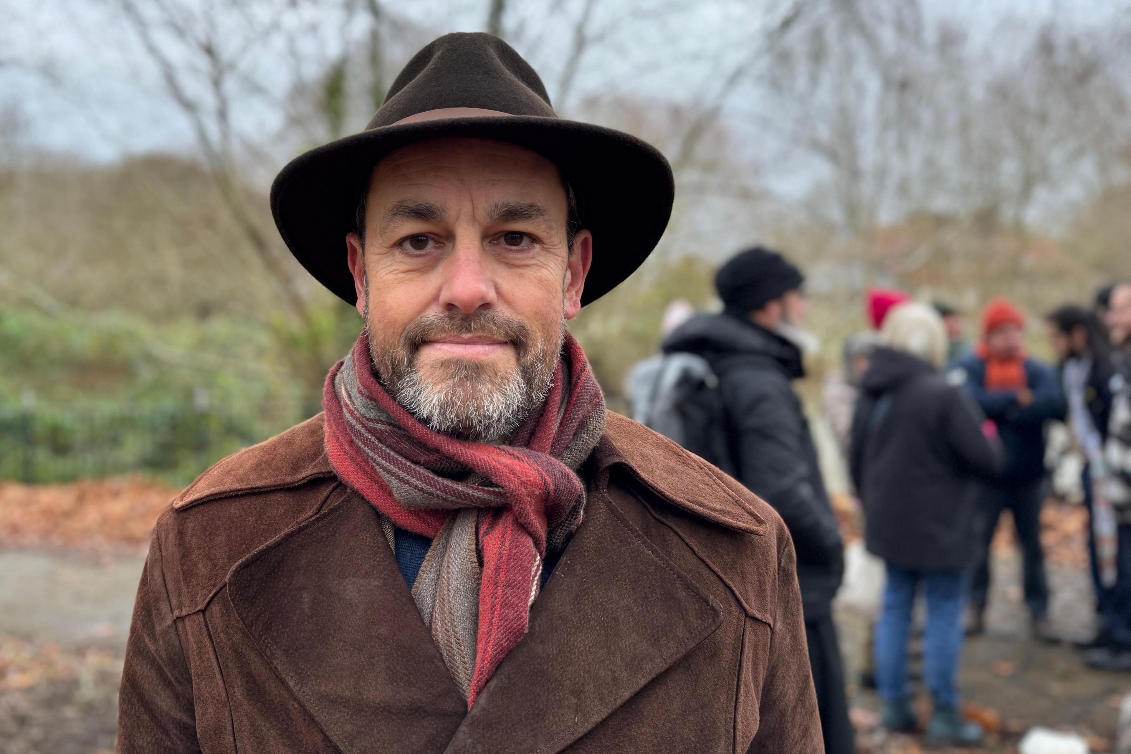 A man in a brown suede or velvet coat, with a striped scarf and trilby hat. Other people are standing behind him in Castle Park. There are trees visible.