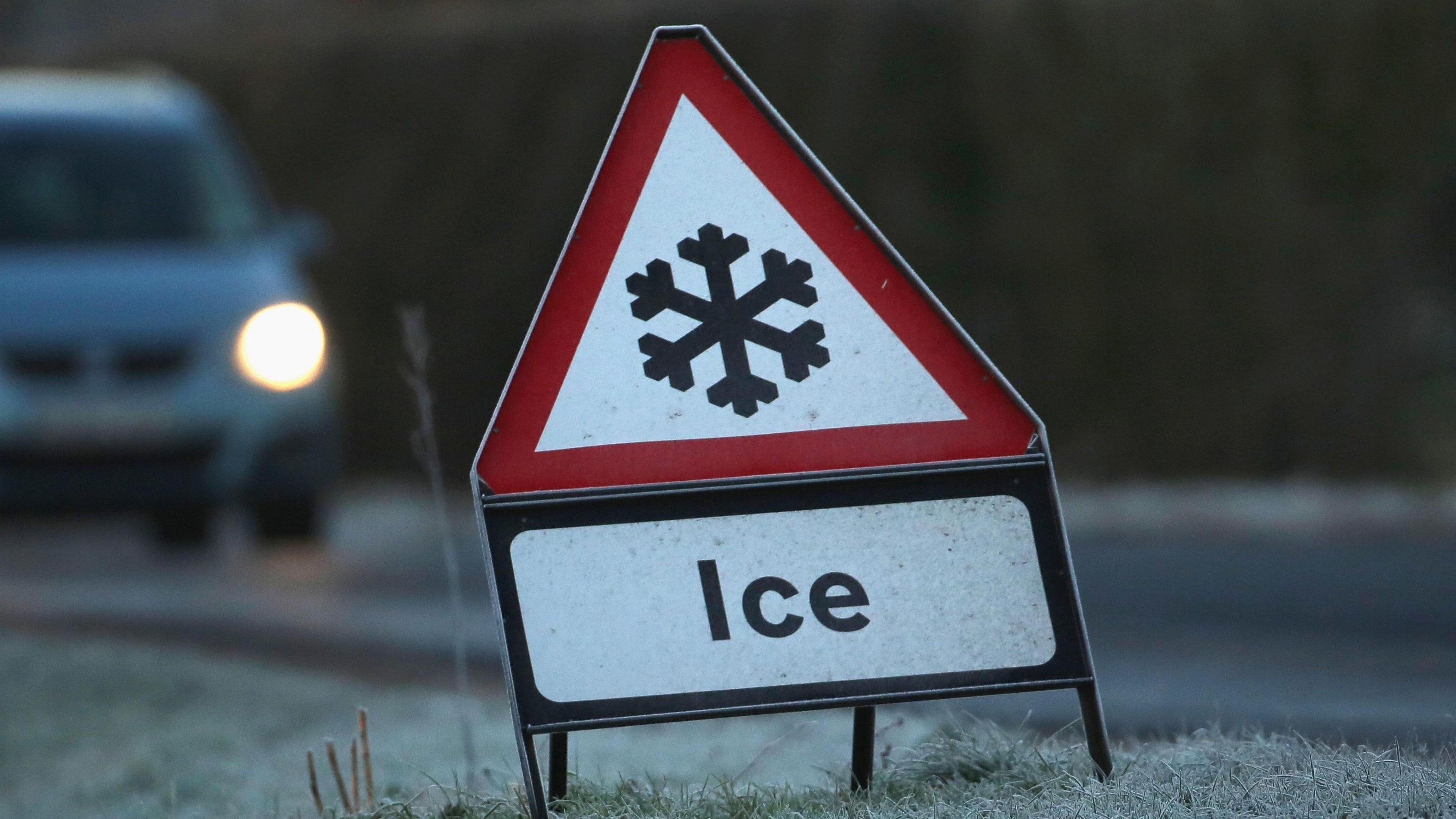 A stock image of a ice warning road sign - a white triangle with a red border showing a black snowflake icon in the middle with Ice written below. The sign is placed on the side of a frosty grass patch. In the background is a blurred silver car driving towards it.