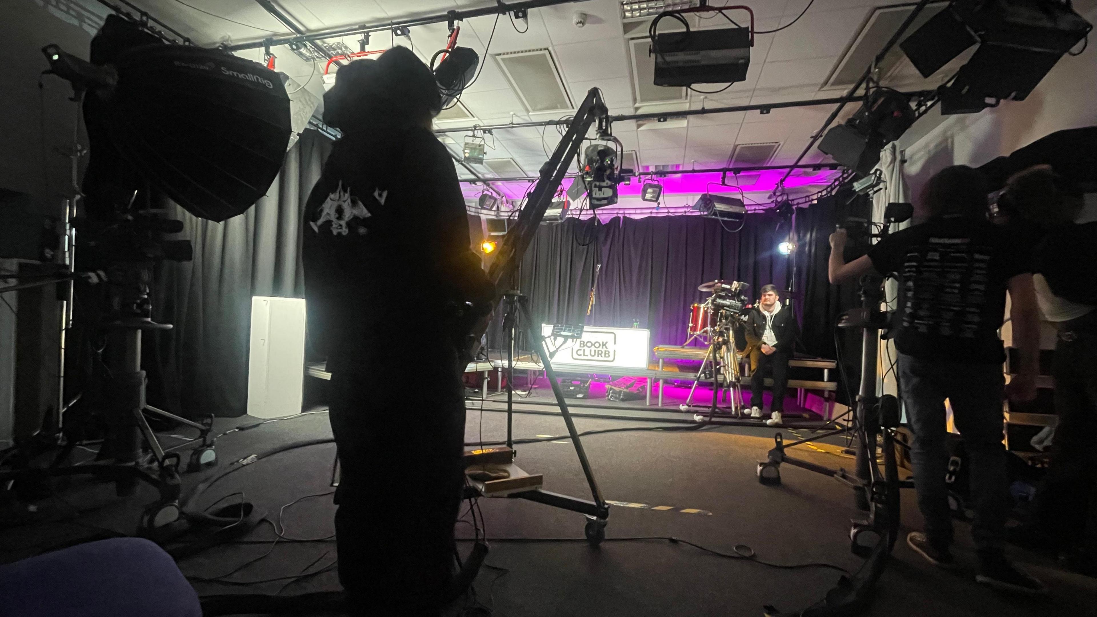 A TV set with lights and cameras pointed to a stage. On stage is a neon sign that reads "The Book Clurb". There is three people stood behind the cameras. 