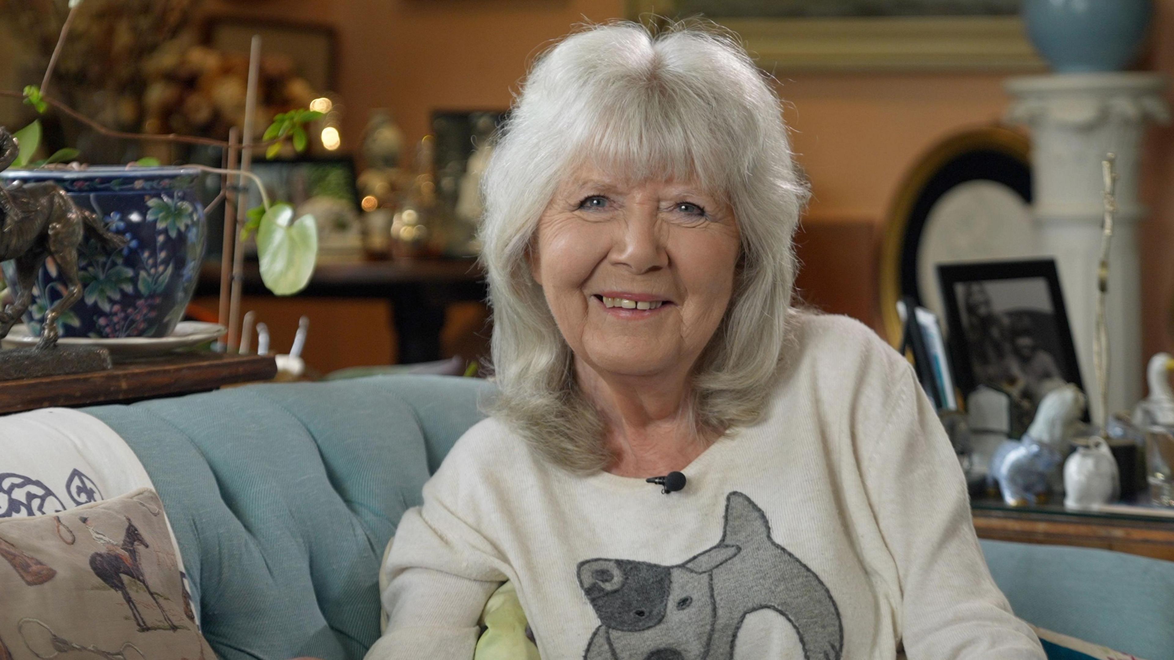 Dame Jilly Cooper sitting on a sofa in the living room of her house in the Cotswolds. She is wearing a dog jumper.