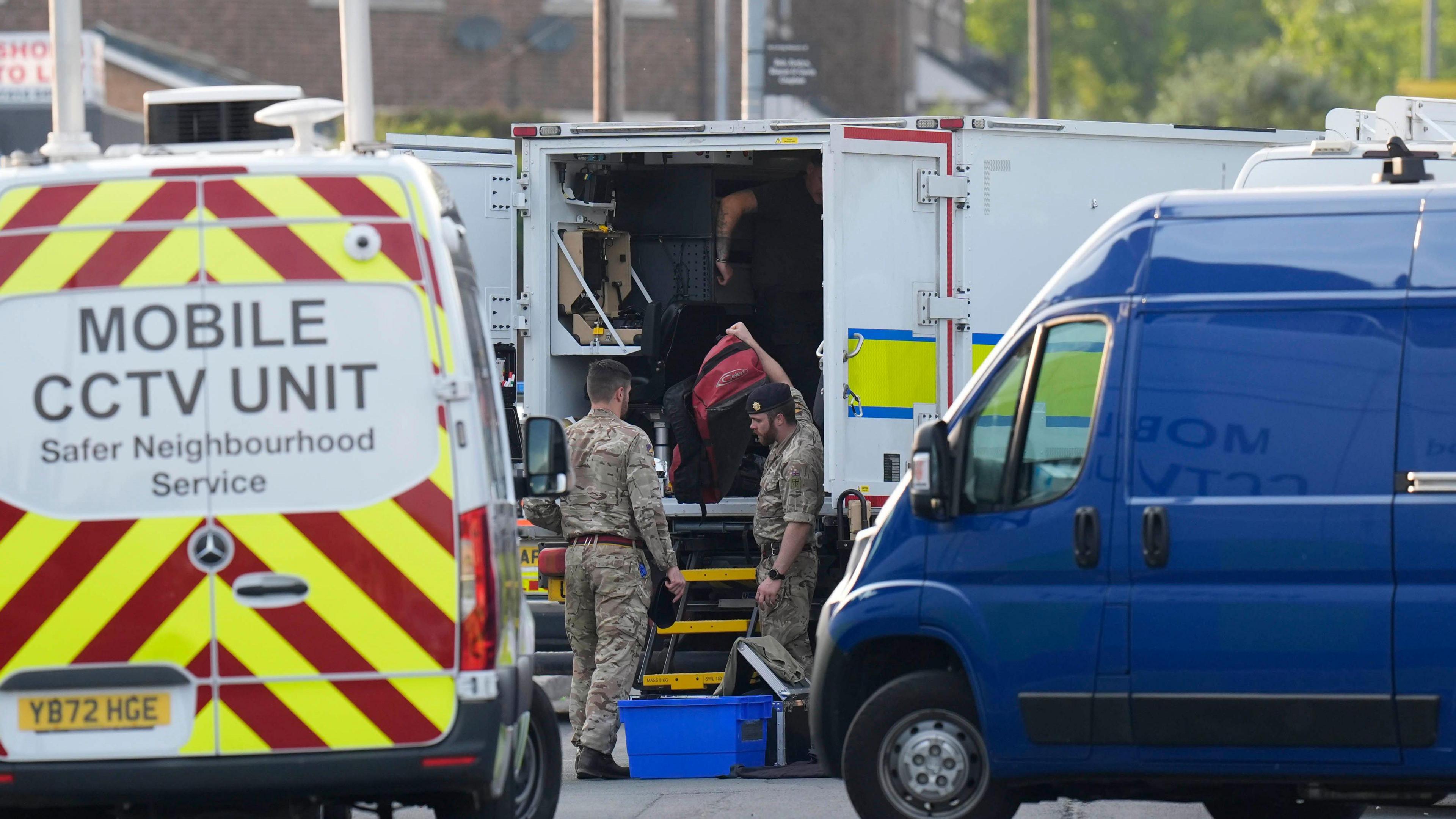 A street scene involving several vehicles and military personnel. In the foreground on the left, there is a white van with bright yellow and red diagonal stripes on the rear and text that reads “MOBILE CCTV UNIT Safer Neighbourhood Service.” To the right, there is a blue van parked nearby. In the centre of the image is a large white vehicle with open rear doors. Two individuals in camouflage uniforms are standing near the open doors, handling equipment, including a blue container on the ground and various gear inside the vehicle. The background shows brick buildings and greenery.