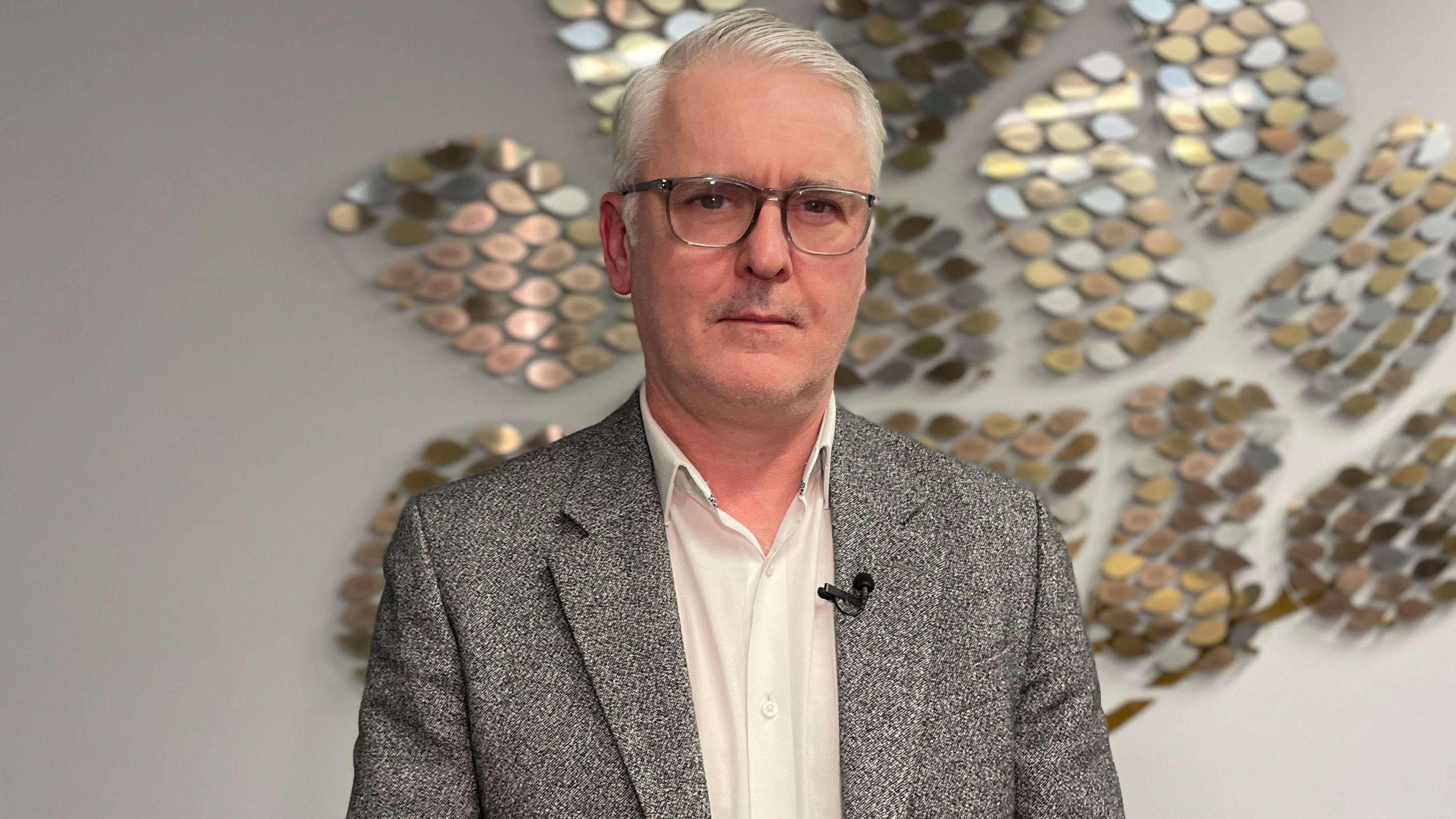 A man with glasses in a grey suit and white shirt stands in front of a metallic artwork