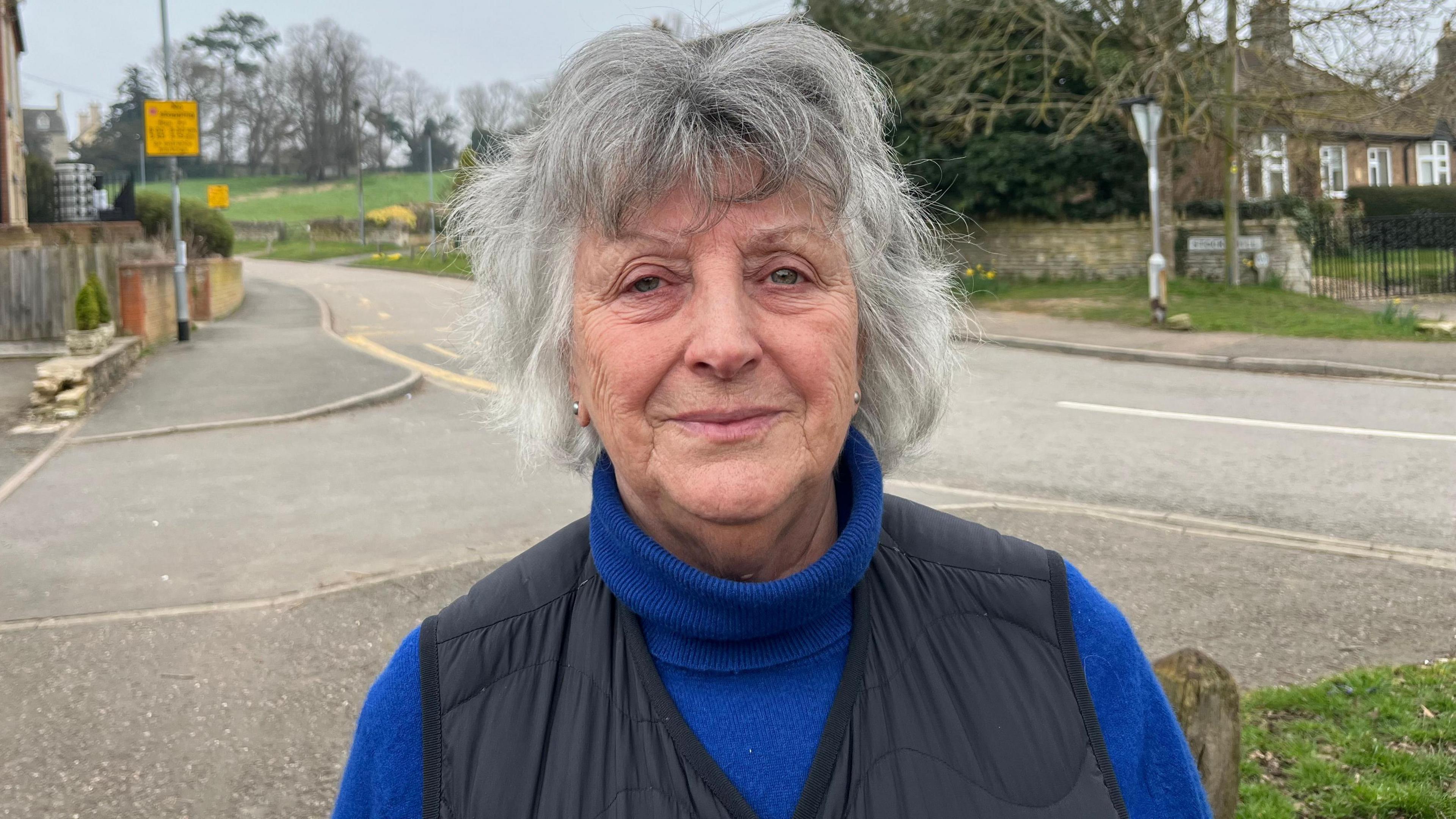 Elaine Wakerley, 73, has short grey hair and is wearing a blue polo neck jumper, with a black body warmer on top. Behind her is a rural road going up a hill, with a grassy field with trees visible behind her. There are properties either side of the road.