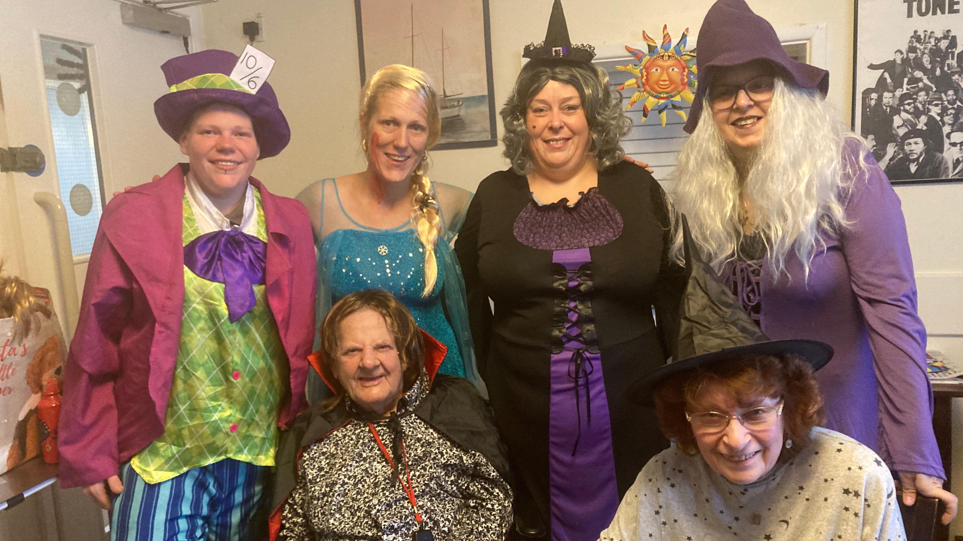 Six women are wearing a range of halloween outfits, including purple and black witches dress with pointy hats, a blue and white sequin dress and a green checkered top and pink blazer 